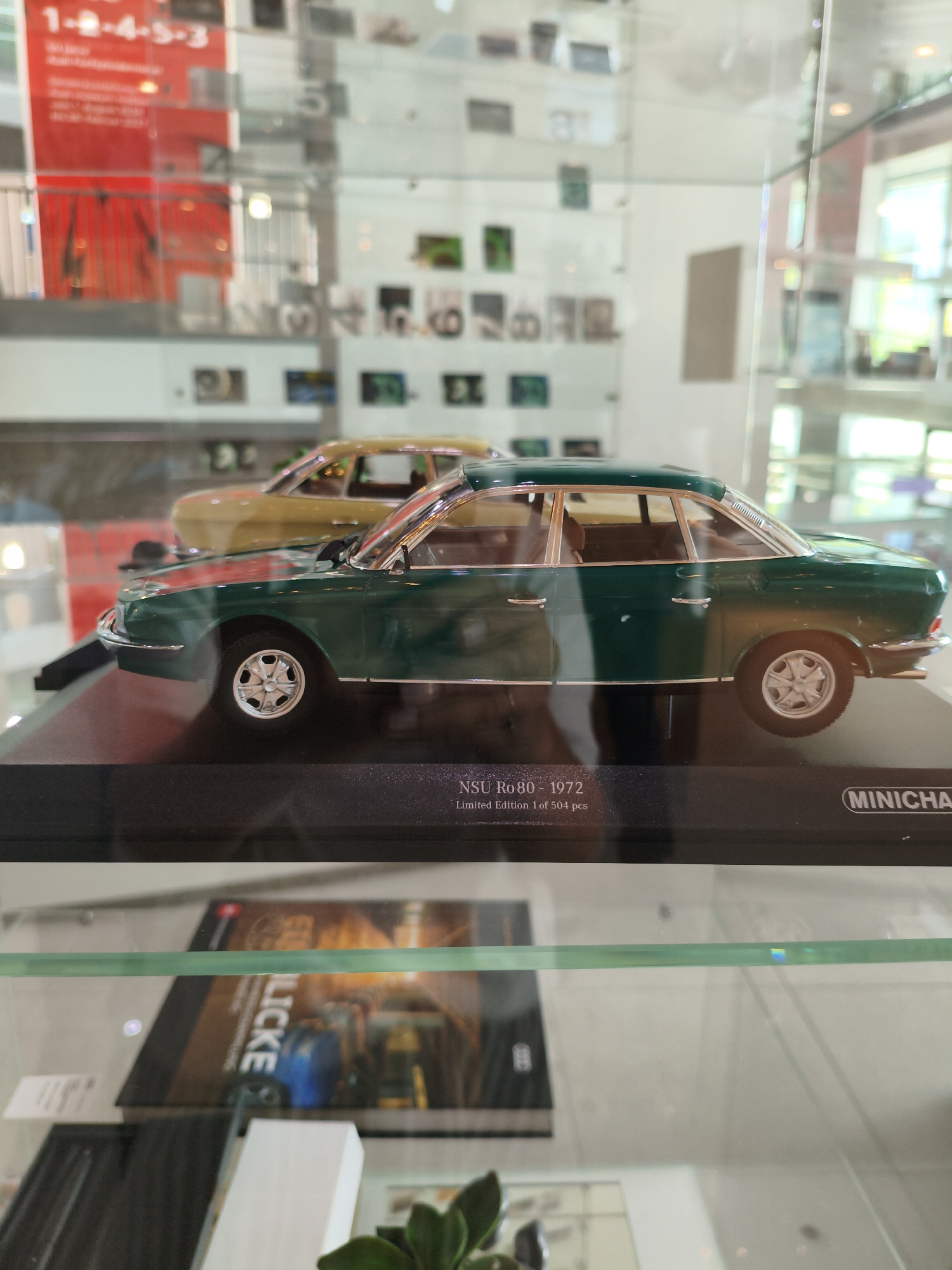 The image captures a display case at the Audi Forum Ingolstadt in Germany. In the foreground, a detailed model of a dark green 1972 NSU Ro 80 sedan is prominently featured on a black display base. The base has text indicating "NSU Ro 80 - 1972" and "Limited Edition 1 of 504 pcs" along with the "MINICH" logo. Behind the green car, a model of a mustard yellow car is partially visible. The display case is made of glass, and reflections are visible on its surface, obscuring some details of the background. The background consists of a white wall adorned with various displays, including framed images and what appear to be informational panels with red and white graphics in the upper left corner. The lighting in the scene is diffused, suggesting an indoor, well-lit environment. Below the display case, a book with the partially visible title "LICKE" is seen. The overall scene suggests a museum or exhibition setting focused on automotive history.