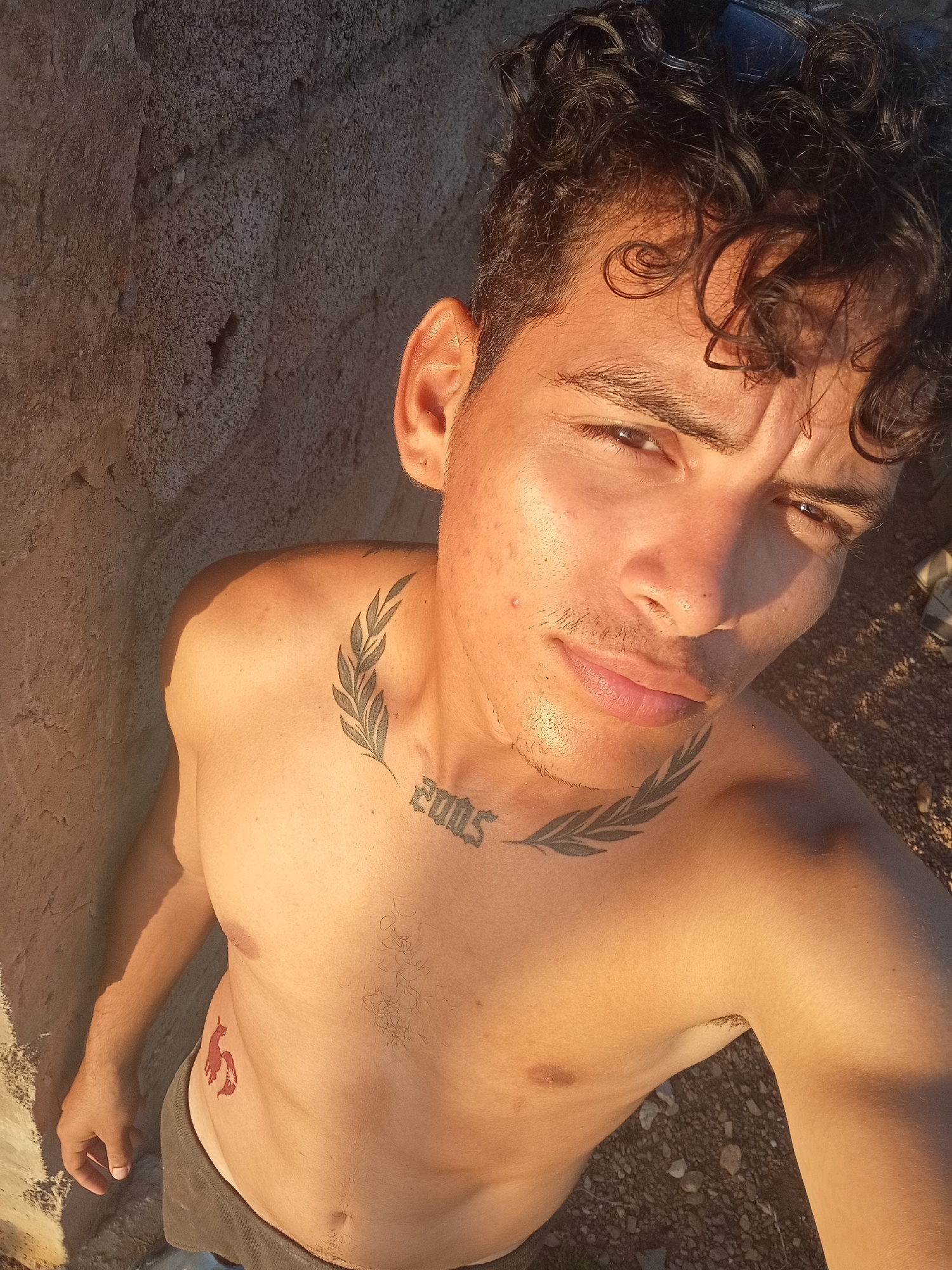 A young adult male with curly dark hair stands near a textured wall in an outdoor environment under bright sunlight. He is shirtless and has a tattoo of the year "2005" with leafy branches on his neck, and another tattoo of a small red animal on his abdomen. He is looking directly at the camera with a slight smirk. The exact location within Altagracia, Venezuela, is not discernible from the image. The background suggests an open space with gravelly ground.