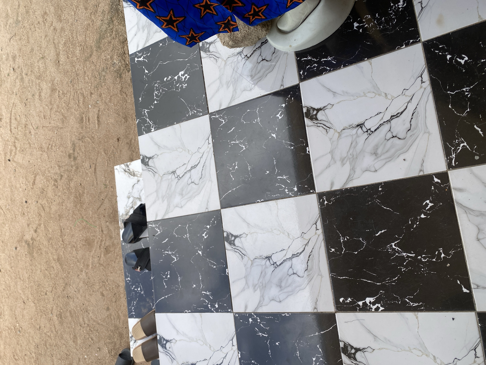 In Jauro Yinu, Nigeria, a pair of brown and black sandals are placed on a tiled step. The step features a checkerboard pattern of black and white marble-effect tiles. To the right, another pair of black sandals rests on the tiles, and a blue and orange patterned fabric is draped over a white mortar and pestle. The background consists of sandy soil.