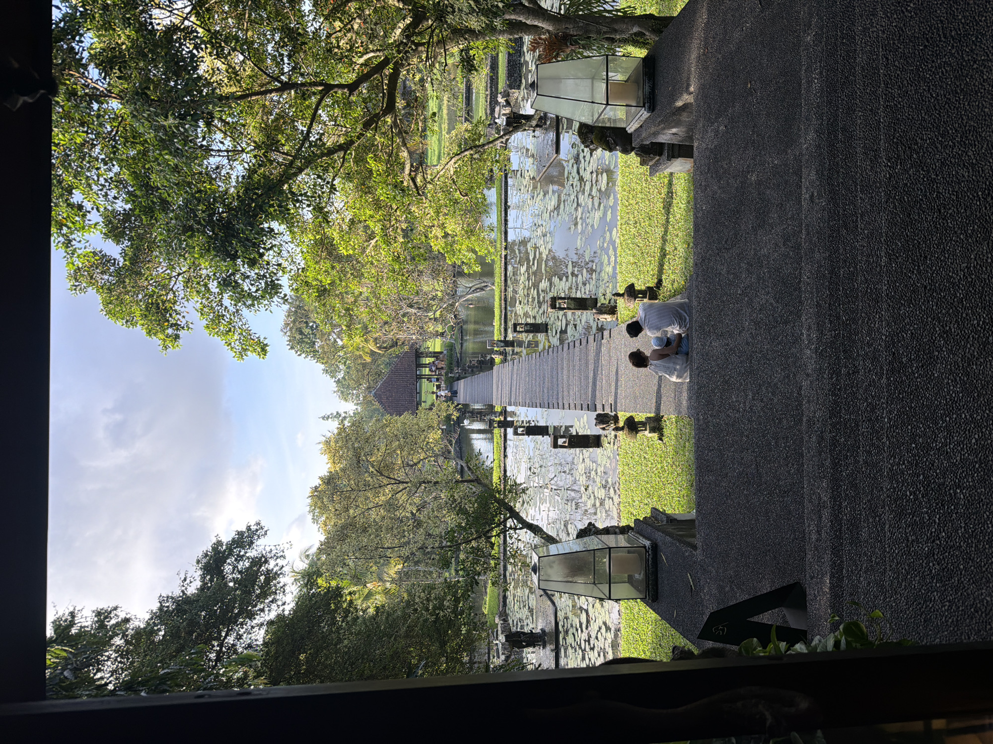The image depicts a view from an elevated position, possibly a building, overlooking a Balinese garden scene, likely in Sukawati, Indonesia. A grey, textured walkway or terrace runs along the right side of the frame, partially obstructing the view. Along the edge of the walkway are black stone structures, some topped with modern, glass-encased lanterns.
Beyond the walkway is a pond or water feature filled with lily pads. A wooden boardwalk extends through the water, leading to a small, traditional Balinese pavilion with a red-tiled roof. People can be seen walking along the boardwalk.
Two people are present closer to the viewer, near the edge of the walkway, appearing to be interacting or observing something in the pond. They are dressed in light-colored clothing.
Lush greenery and trees surround the water feature, creating a tranquil environment. The sky is partly cloudy, suggesting it is daytime. The image is framed by a window or architectural structure, partially visible at the bottom and left edge of the frame.