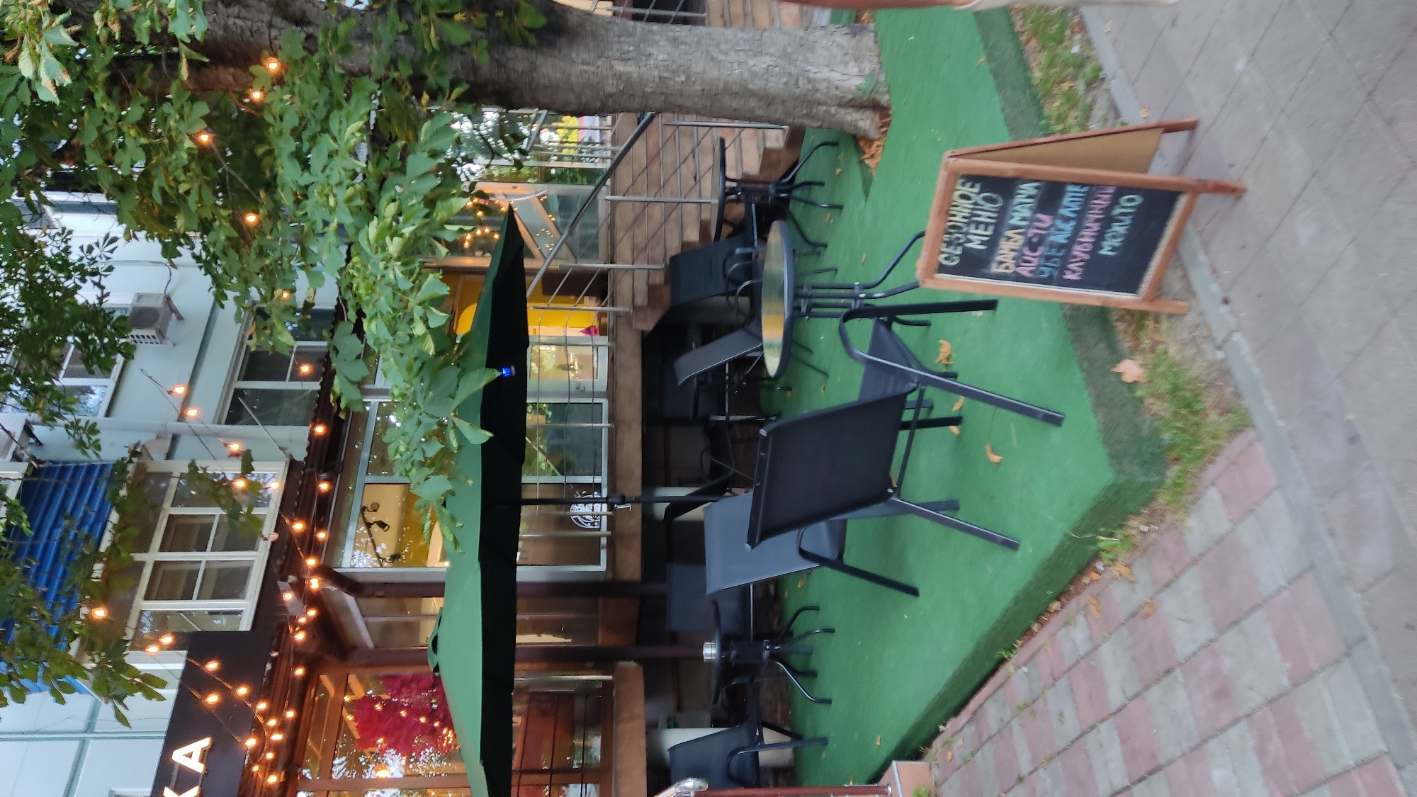 The image depicts an outdoor seating area of a cafe, likely in Krasnodar, Russia, given the Cyrillic text on the menu board. The cafe features green artificial turf flooring and black metal tables and chairs. A large green umbrella provides shade. String lights are draped across the building's facade, which has large windows. The building appears to be a multi-story structure with light-colored walls. A tree with green leaves stands to the right of the seating area, and stairs are visible behind the tables. A blackboard menu, with Cyrillic writing, is placed on the pavement in the foreground, listing seasonal menu items including "БАМБА МАТЧА" (BAMBA MATCHA), "AUC-TU", "УБЕ АС АМПТЕ" (UBE AS AMPTE), "КЛУБИЧНИЧНЫЙ" (STRAWBERRY), and "МОХИТО" (MOJITO). The overall scene suggests a pleasant outdoor dining or relaxation space.