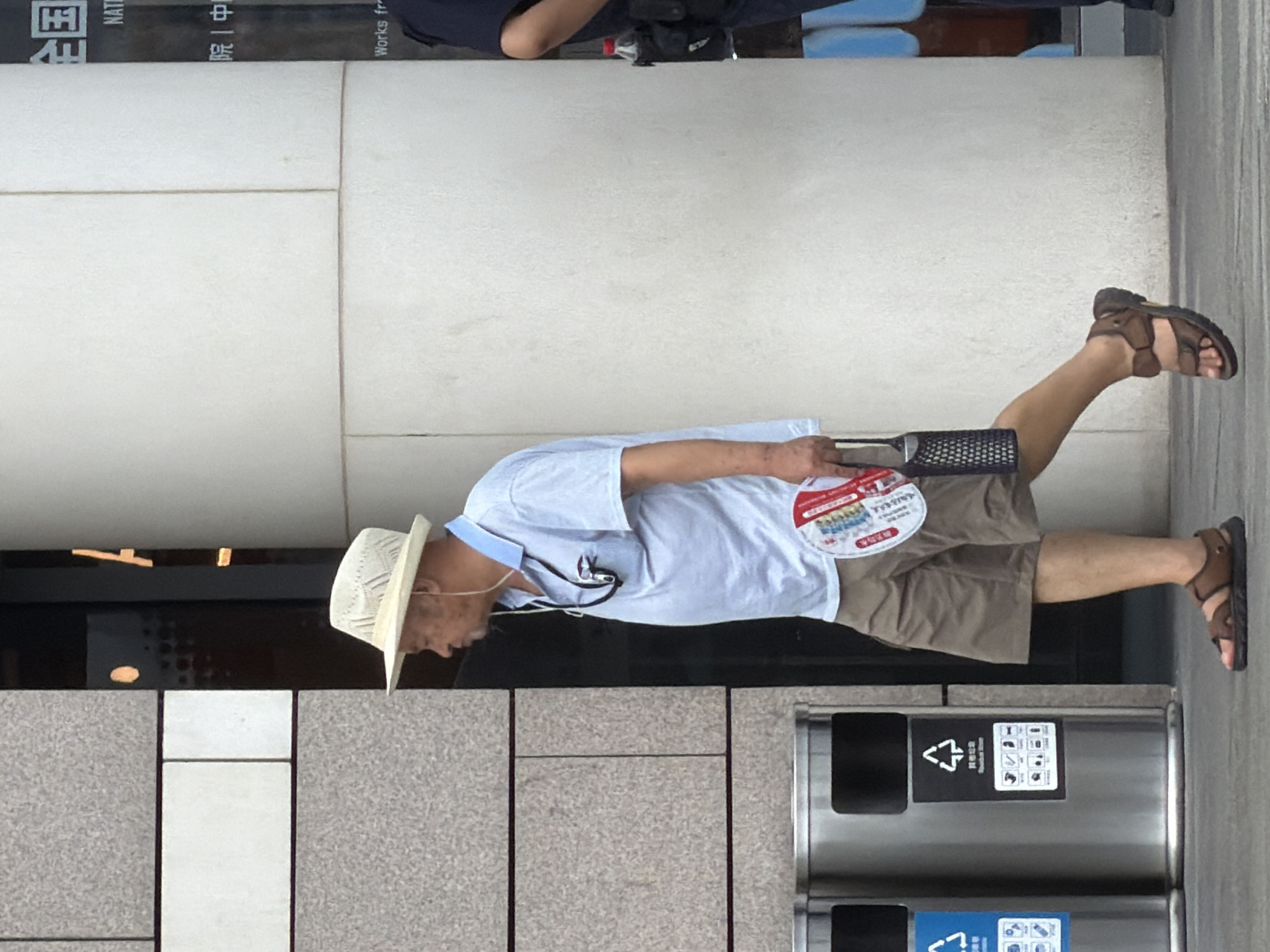 The image shows an elderly East Asian man wearing a light-colored woven hat, a pale blue polo shirt, and khaki shorts. He is walking from left to right in the frame, holding a round red and white fan and a black cylindrical object in his right hand. He is also wearing brown sandals. The man is walking on a paved sidewalk next to a large, textured concrete pillar on his right. To his left, there are two cylindrical silver trash bins. In the background, a building with a grey stone facade is visible on the far left, and a section of the concrete pillar has text on it, including "NATIONAL" and "WORKS fr". The scene appears to be in a city environment, likely Yuzhong District, China, given the context provided. Another person, partially visible on the far right, is wearing a dark uniform, suggesting a public or security role. The lighting indicates it is daytime.