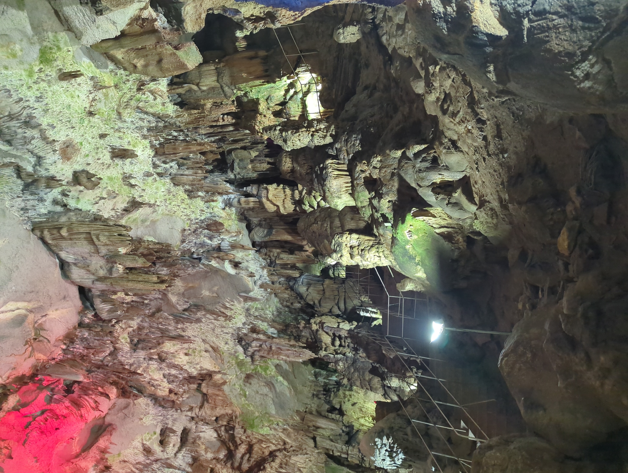 The image depicts the interior of a cave, likely a tourist attraction due to the presence of a metal staircase with handrails and a spotlight illuminating the path. Stalactites and stalagmites are abundant, forming intricate formations on the cave's ceiling and floor. Some formations are illuminated by colored lights, with a prominent red glow on the upper left and a greenish hue on the upper right. The cave walls are rugged and textured, composed of rock formations. The ground is uneven, covered with rocks and debris. The lighting creates a dramatic effect, highlighting the natural beauty of the cave's geological features. This scene is consistent with the appearance of the Vratsa cave in Vratsa, Bulgaria, which is known for its accessible passages and illuminated formations.
