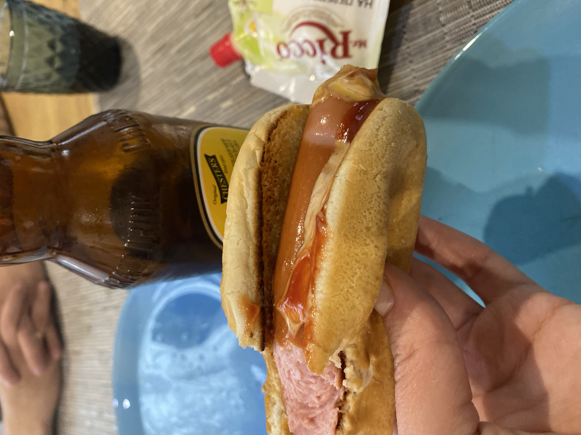 A close-up shot shows a hand holding a hot dog in a bun, partially eaten, with ketchup drizzled over it. In the background, a brown glass bottle with a yellow label that reads "CHESTER'S" is visible, along with a small packet of "Ricco" sauce. A blue plate and a green glass are also in the scene, all placed on a table with a textured gray placemat. The lighting suggests an indoor setting.