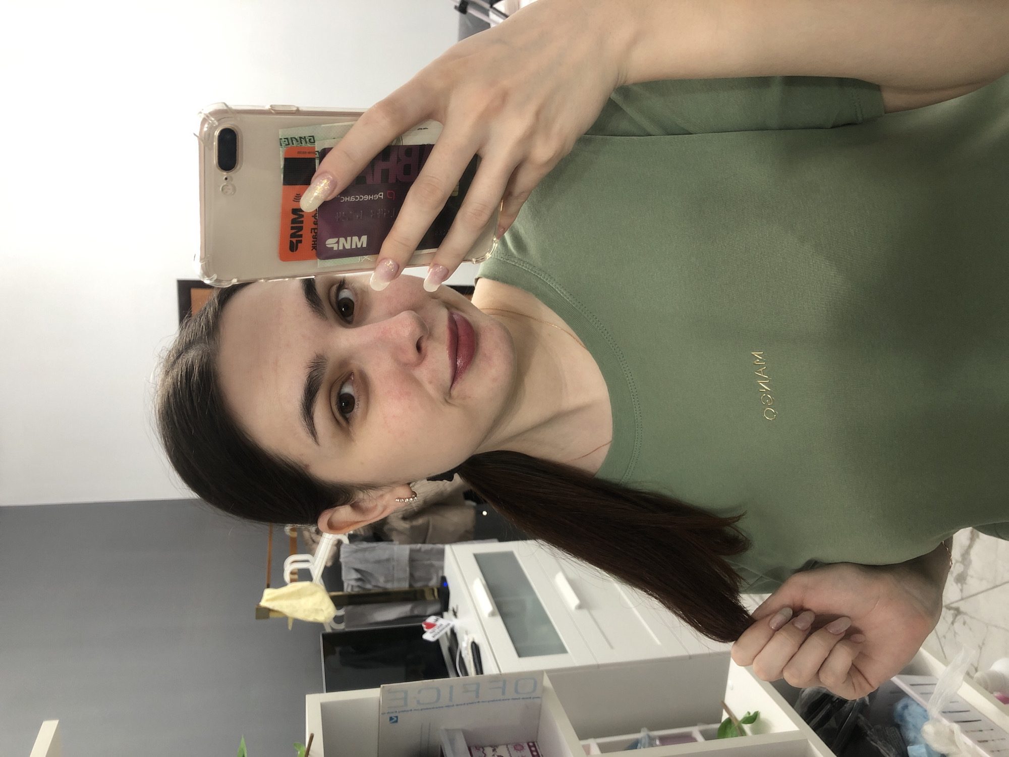 A woman takes a selfie in what appears to be an office or study space in an indoor environment. The woman is facing the camera, and she is holding a mobile phone in front of her face to capture the picture. Her nails are long, with a light-colored base and glittering tips. The phone has credit cards in the back. She is wearing a light green t-shirt with the word "MANGO" visible on the front.
In the background, there is a white shelving unit with various items. Part of a computer screen, a white cabinet and a clothes rack are also visible. There is no indication of the exact location in the city of Chaikovsky, Russia.