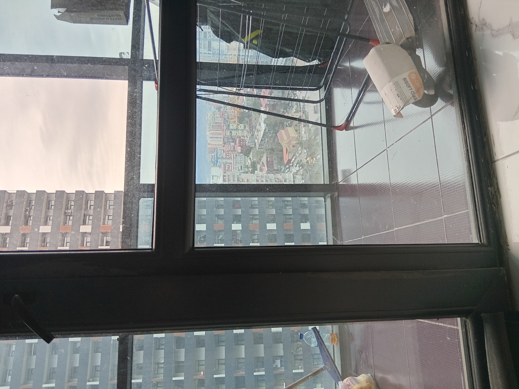 A view through a window reveals a cityscape, likely Batumi, Georgia. High-rise buildings with repeating window patterns dominate the view, contrasting with colorful residential structures visible in the mid-distance. A clothes drying rack and cleaning supplies are visible within the indoor setting. The window frame is dark and partially obstructs the view. The lighting suggests an overcast day.
