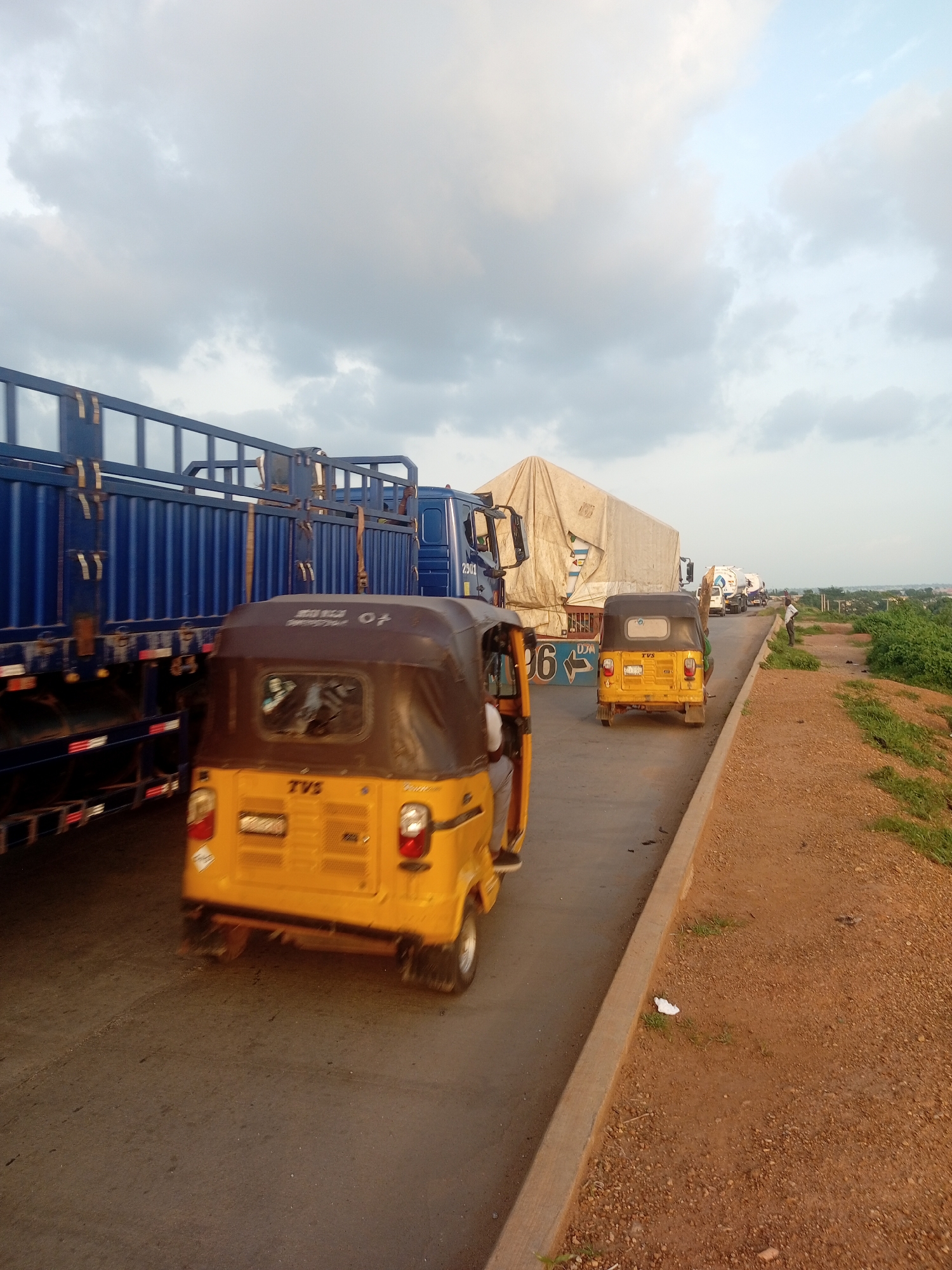 The image depicts a traffic jam on a paved road under a cloudy sky, likely during the late afternoon or early evening. The scene is dominated by various vehicles, including large blue trucks, a white-sheet-covered truck, and several yellow auto-rickshaws (also known as tuk-tuks). One auto-rickshaw in the foreground is clearly visible, showing the "TVS" logo on its rear. Another auto-rickshaw is positioned behind it, and further down the road, more vehicles are seen in the congestion. The road is bordered on the right by a dirt embankment with sparse vegetation. There are hints of human presence with a person visible in the driver's seat of the foreground auto-rickshaw and a few figures standing further down the road near the embankment. The overall mood is one of stagnation and waiting, characteristic of a traffic standstill. The lighting suggests the sun is low in the sky, casting a soft, diffused light through the clouds.