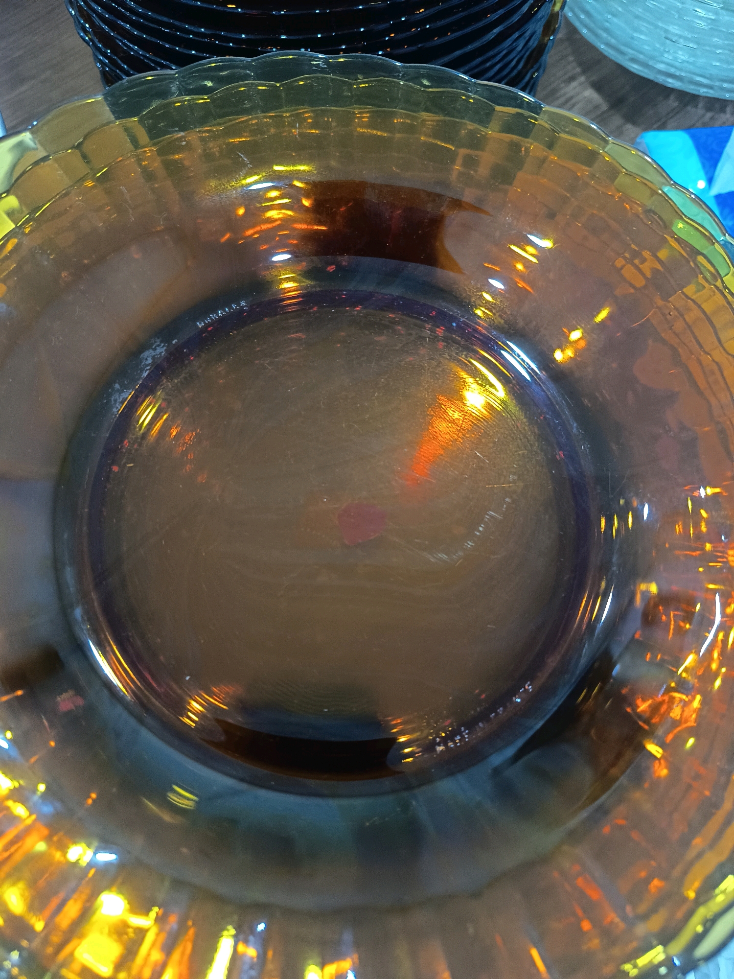 The image shows a close-up, overhead view of a stack of amber-colored glass plates with scalloped edges. The plates are reflecting light, creating a warm, glowing effect. In the background, other stacks of dishes, including dark blue ones and clear ones, are visible. The setting appears to be indoors, possibly a kitchen or dining area. The image does not contain any elements that would allow for the determination of its location within the city of Bogor, Indonesia.