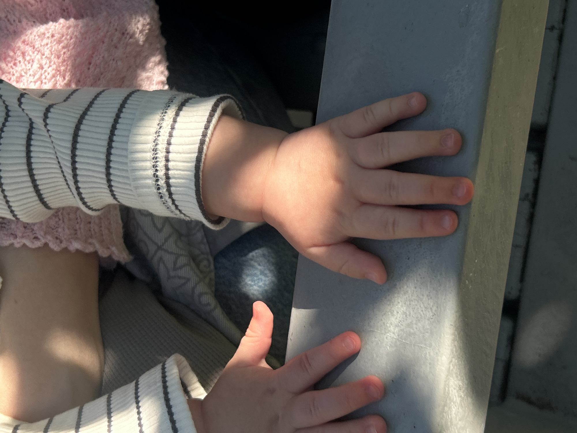 The image shows a close-up of a child's hands and arms, resting on a grey surface. The child is wearing a white long-sleeved shirt with black stripes and a pink knitted garment. Sunlight casts a shadow across the child's right hand and arm. The background is blurred, showing a hint of patterned fabric. The scene appears to be outdoors, possibly in a park or garden. There is no specific indication of a location in Saint Petersburg, Russia, within the image itself.