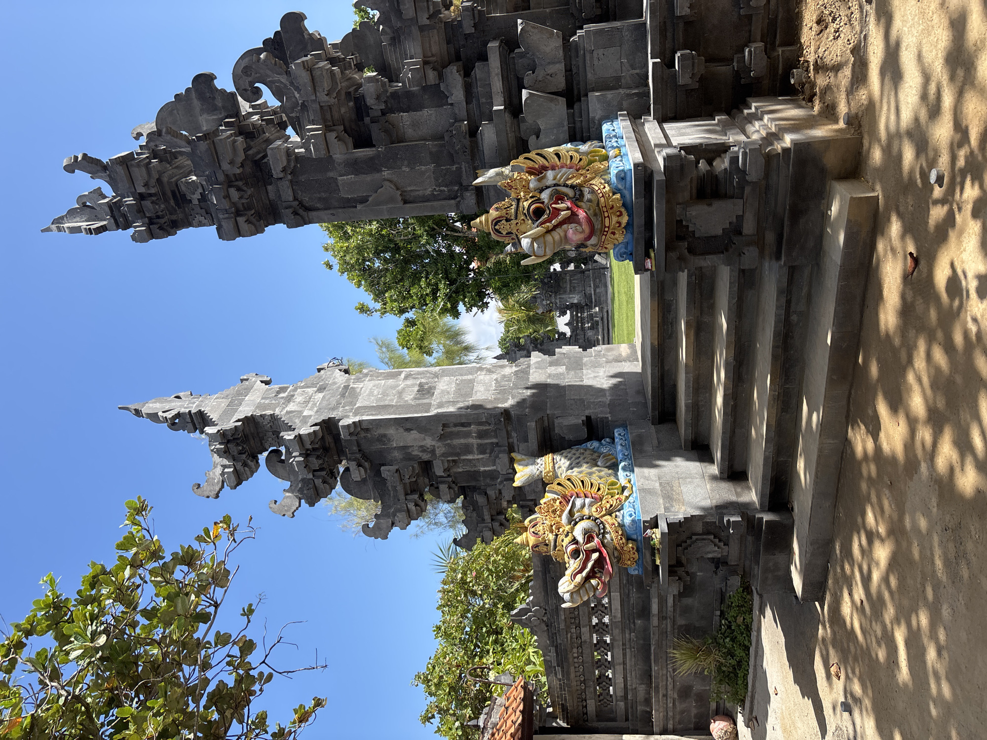 In Kuta Selatan, Indonesia, a stone gateway stands under a clear blue sky. The gateway is flanked by two colorful statues of mythical creatures with dragon-like heads and golden headdresses. Steps lead up to the gateway, which is adorned with intricate carvings. Lush green trees and foliage surround the structure, creating a vibrant contrast with the dark stone. The ground in front of the gateway is covered with sand and fallen leaves, and dappled sunlight filters through the trees, casting shadows on the ground.