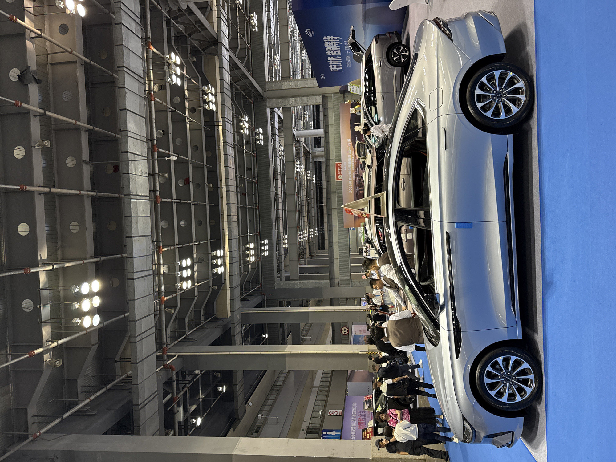 The image shows a car exhibition hall, possibly in Chongqing, China, based on the Chinese writing on the banners. The hall is filled with people looking at various cars. The architecture features high ceilings with exposed metal beams and numerous lights. In the foreground, there is a silver car with its door open, surrounded by people. The floor is covered with a blue mat.