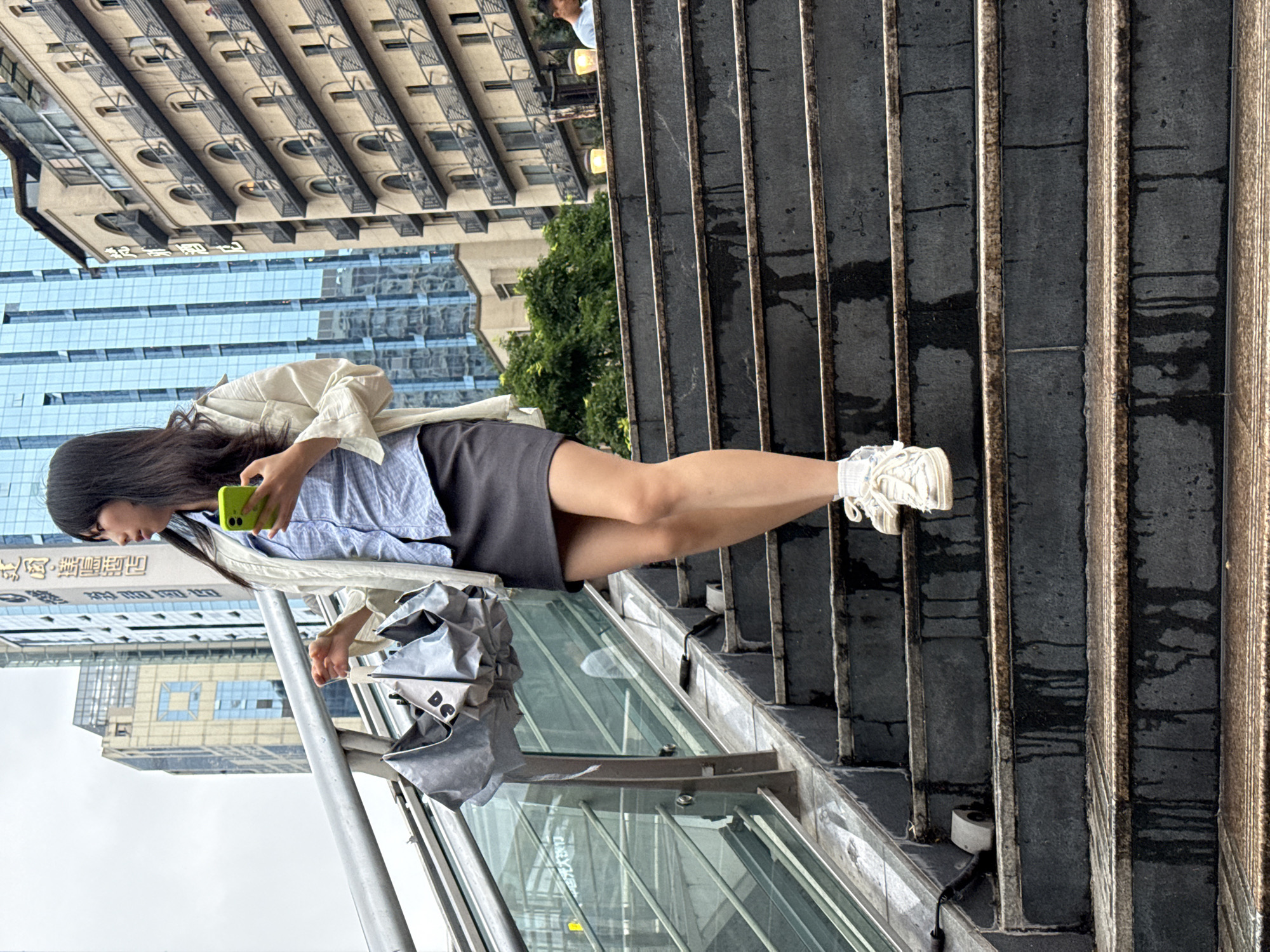 In Yuzhong District, China, a young Asian woman with long black hair walks down a wet, dark grey outdoor staircase. She is wearing a light-colored jacket over a blue collared shirt and a dark grey skirt. She is holding a folded grey umbrella in her left hand and a bright green smartphone in her right hand. Her white sneakers are visible as she descends the stairs. The background shows modern buildings, including a tall blue glass skyscraper and a beige building with arched balconies. The overall atmosphere suggests a cloudy day in an urban environment.