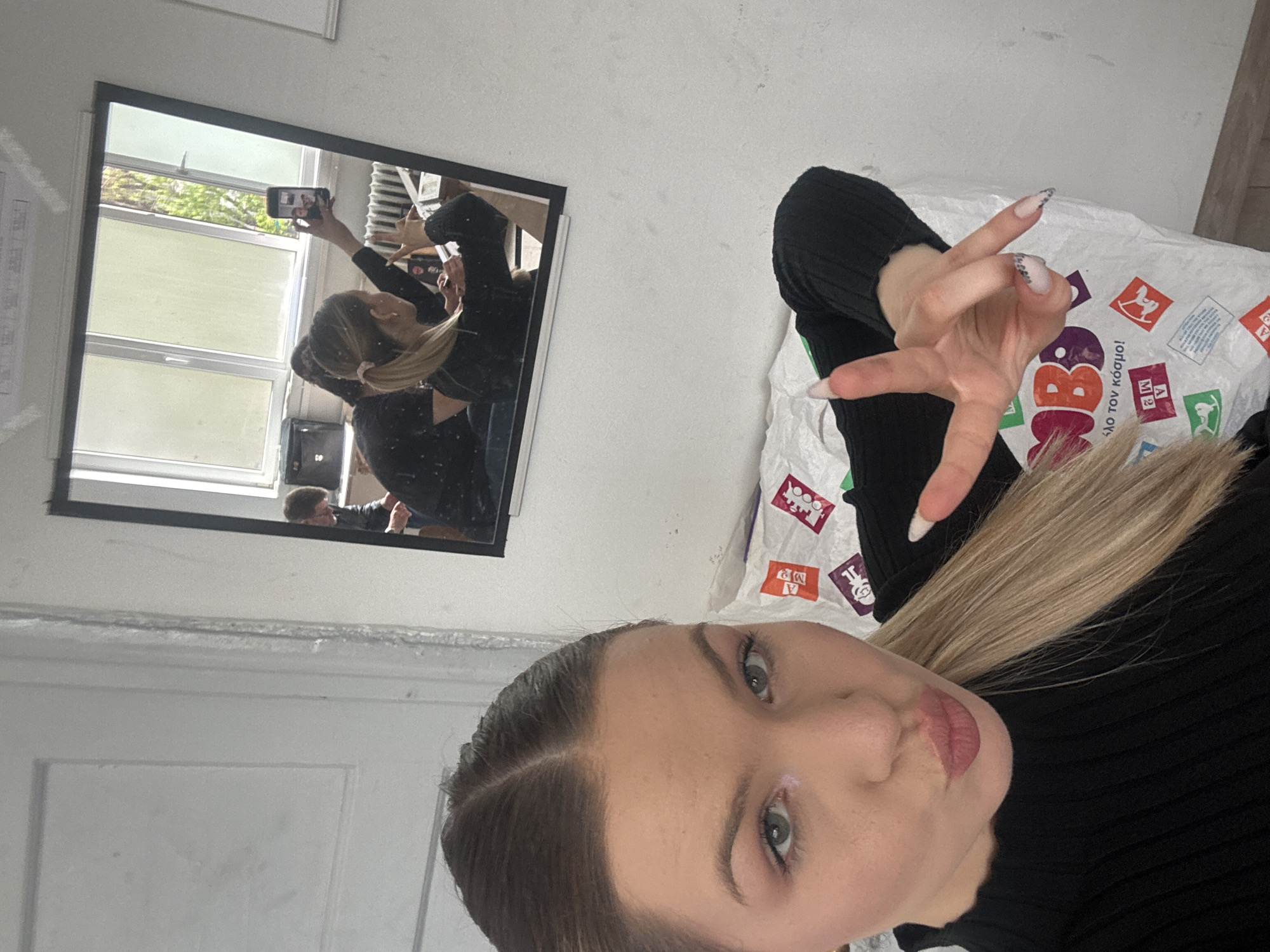 A young woman is captured taking a mirror selfie indoors, likely in Plovdiv, Bulgaria, given the visible Greek text. She is seen from the chest up, wearing a black ribbed long-sleeved top. Her long blonde hair is partially visible, and she has long, light-colored nails with a subtle pattern. Her right hand is raised, making a gesture with spread fingers. She has a calm expression with slightly pursed lips, looking towards the camera (which is her phone).
Her reflection in the large rectangular mirror shows her holding a black smartphone up to capture the image. In the mirror's background, a window reveals a daytime scene with greenery outside, indicating bright conditions. Another person, a male, is faintly visible in the lower part of the mirror's reflection.
To the right of the woman, a white, plastic-like bag or covering is visible, adorned with colorful squares containing fragmented Greek text and various symbols. Clearly legible words include "ΒΑΘΜΟΙ" (VATHMOI, meaning 'degrees' or 'grades') and "σάλο τον κόσμο!" (salo ton kosmo, meaning 'the world's craze' or 'shake the world!'). The room features white walls, and a light-colored floor is partially visible on the right.
