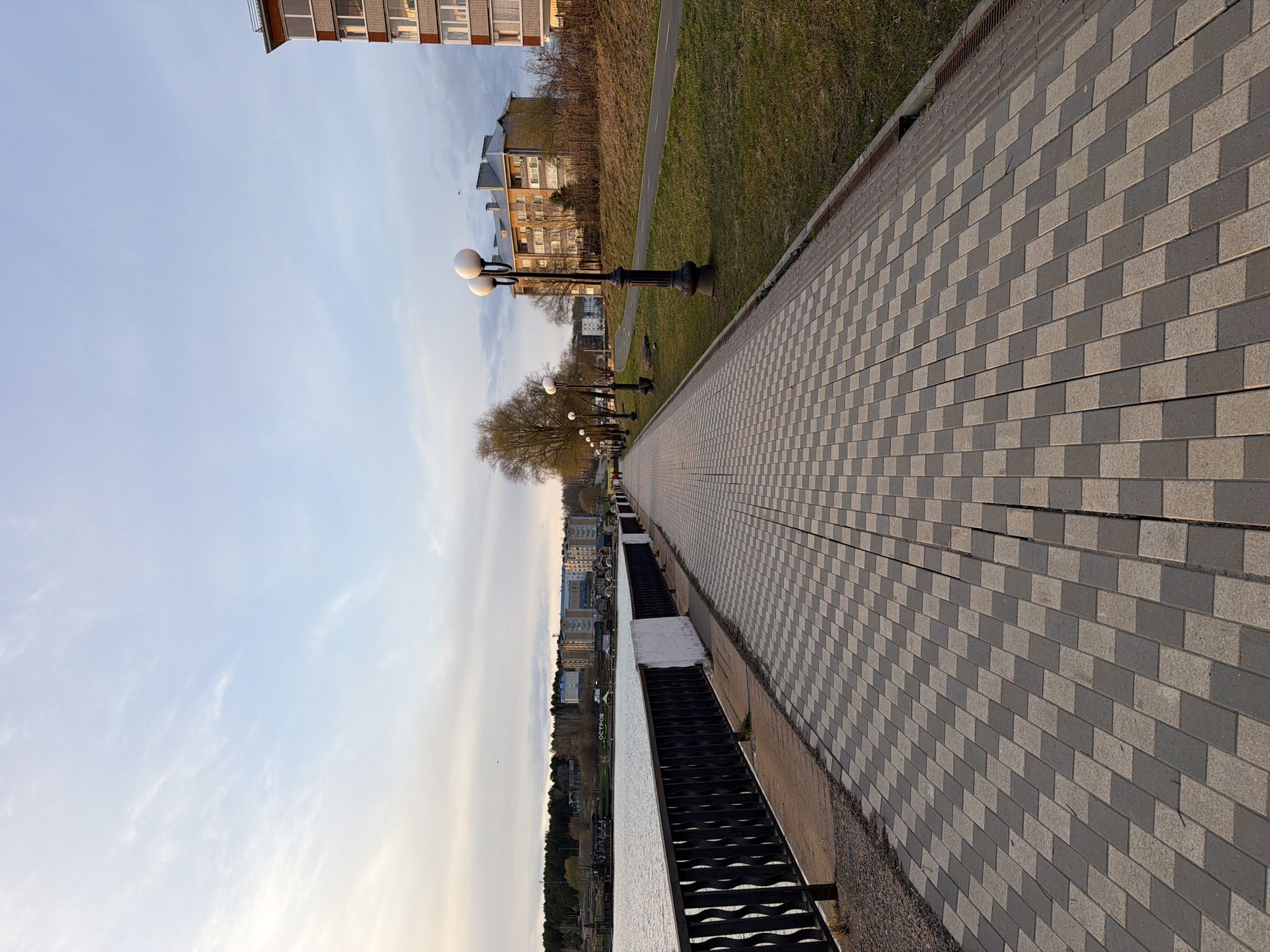 The image depicts a street scene in Votkinsk, Russia. A wide pedestrian walkway paved with gray and light gray checkered bricks dominates the right side of the image. The walkway is bordered by a strip of grass and then a road with some buildings and trees in the background. On the left side of the image, there's a promenade with black railings. Beyond the railings is a body of water. In the distance, there are buildings on the opposite bank. There are black lampposts along the walkway. The sky is light blue with some clouds. The overall perspective suggests a low angle or wide-angle lens.