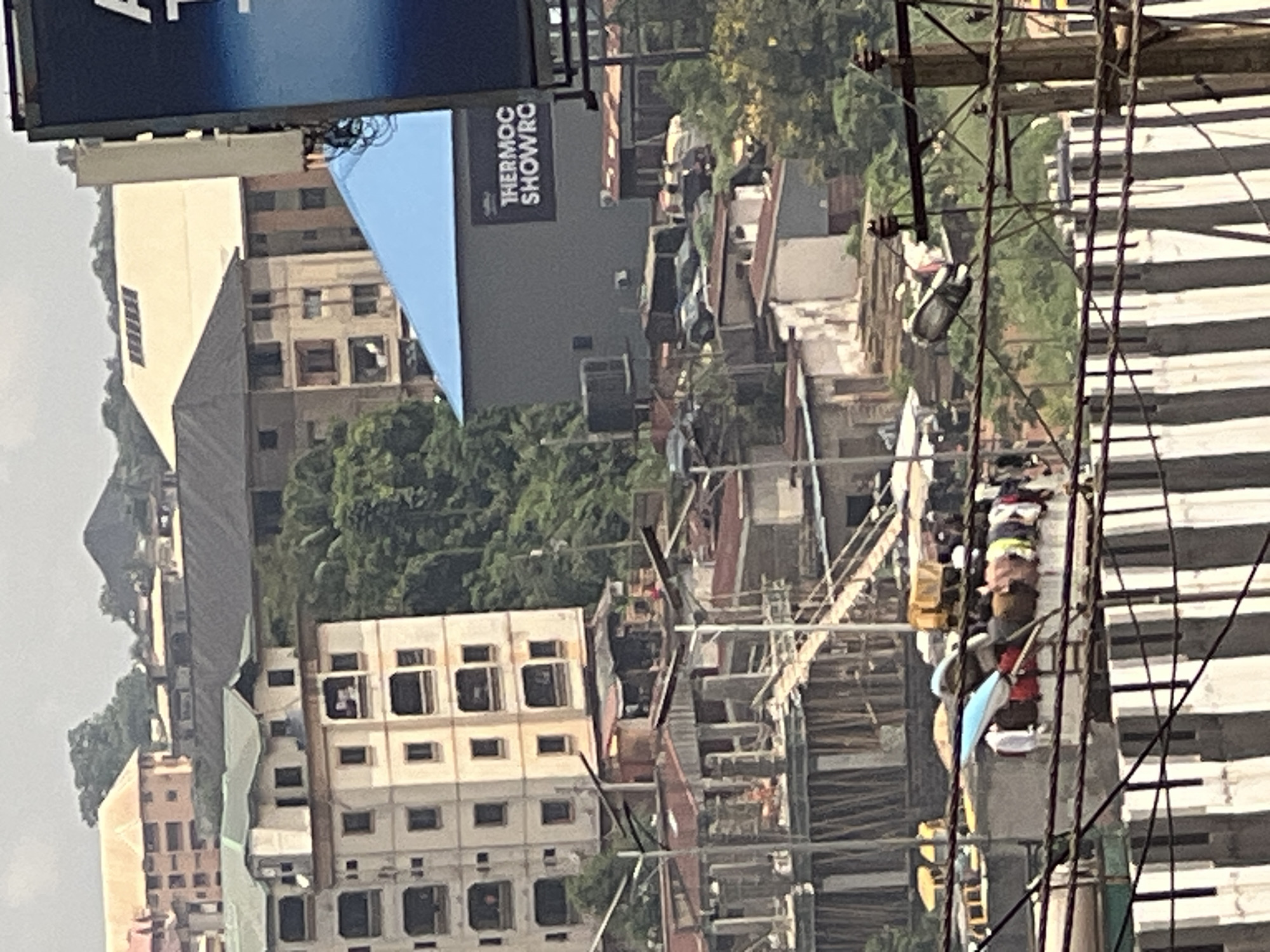 This image depicts a bustling urban landscape in Nkpor, Nigeria, characterized by a dense collection of buildings, dense vegetation, and various modes of transportation. In the foreground, a series of wires and a concrete barrier suggest an elevated vantage point or a highway overpass. Beyond this, a construction site is visible with scaffolding and building materials, indicating ongoing development.
Dominating the mid-ground are multi-story buildings, some appearing residential with balconies, others commercial with signage that reads "THERMOC SHOWRO". These structures are interspersed with lush green trees, suggesting a tropical environment. Vehicles, including cars and what appear to be yellow vans or buses, are visible on the streets, indicating active traffic. A section of the image shows a clothesline with laundry hanging to dry, a common sight in residential areas.
In the background, more buildings of varying architectural styles are spread across rolling hills, with some structures seemingly built into the hillsides. The sky is overcast, suggesting a cloudy day. The overall impression is one of a vibrant, densely populated city with a mix of modern development and older structures, all set against a backdrop of natural greenery.