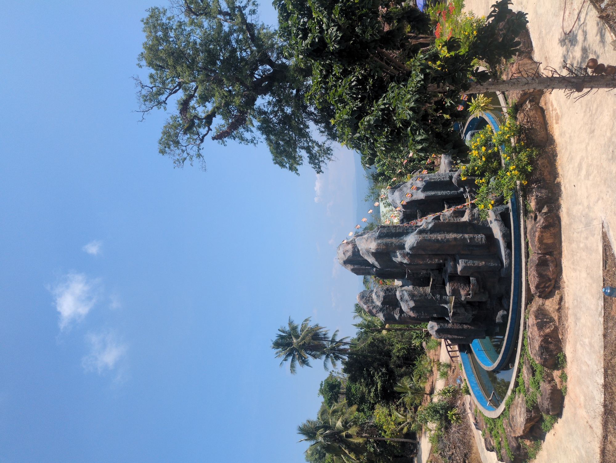 The image depicts a water park attraction, featuring a rock structure with built-in pools or channels, situated within a tropical environment. Palm trees and other vegetation surround the attraction. The clear blue sky suggests a sunny day. The location is the city of Phú Quốc, Vietnam. The rock structure is decorated with small flags or banners. The pools contain blue water. The ground around the attraction is paved. The angle of the photograph suggests it was taken from an elevated position.