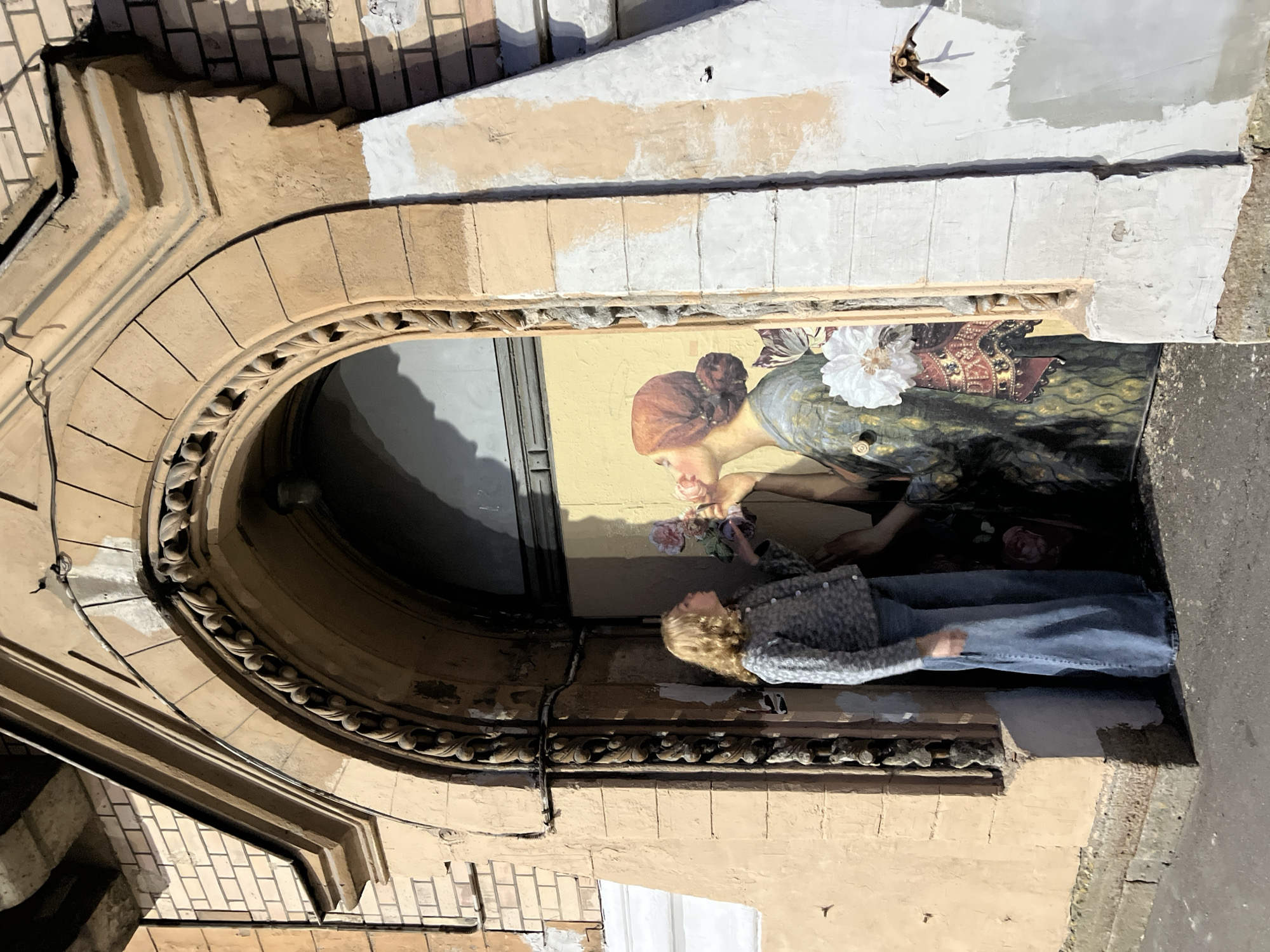 The image depicts a young woman standing in front of a mural on a building in Saint Petersburg, Russia. The woman, with blonde, curly hair, is wearing a grey patterned cardigan and wide-leg blue jeans. She is reaching out to a flower depicted in the mural. The mural features a woman with her hair in a bun, wearing a dark, ornate dress and kissing a pink flower. The mural is painted on a yellow surface within an arched doorway of an old, light-peach colored building. The building's facade shows signs of wear and repair, with some areas painted white. The doorway has decorative stone carvings around the arch. The ground in front of the building appears to be a paved street.