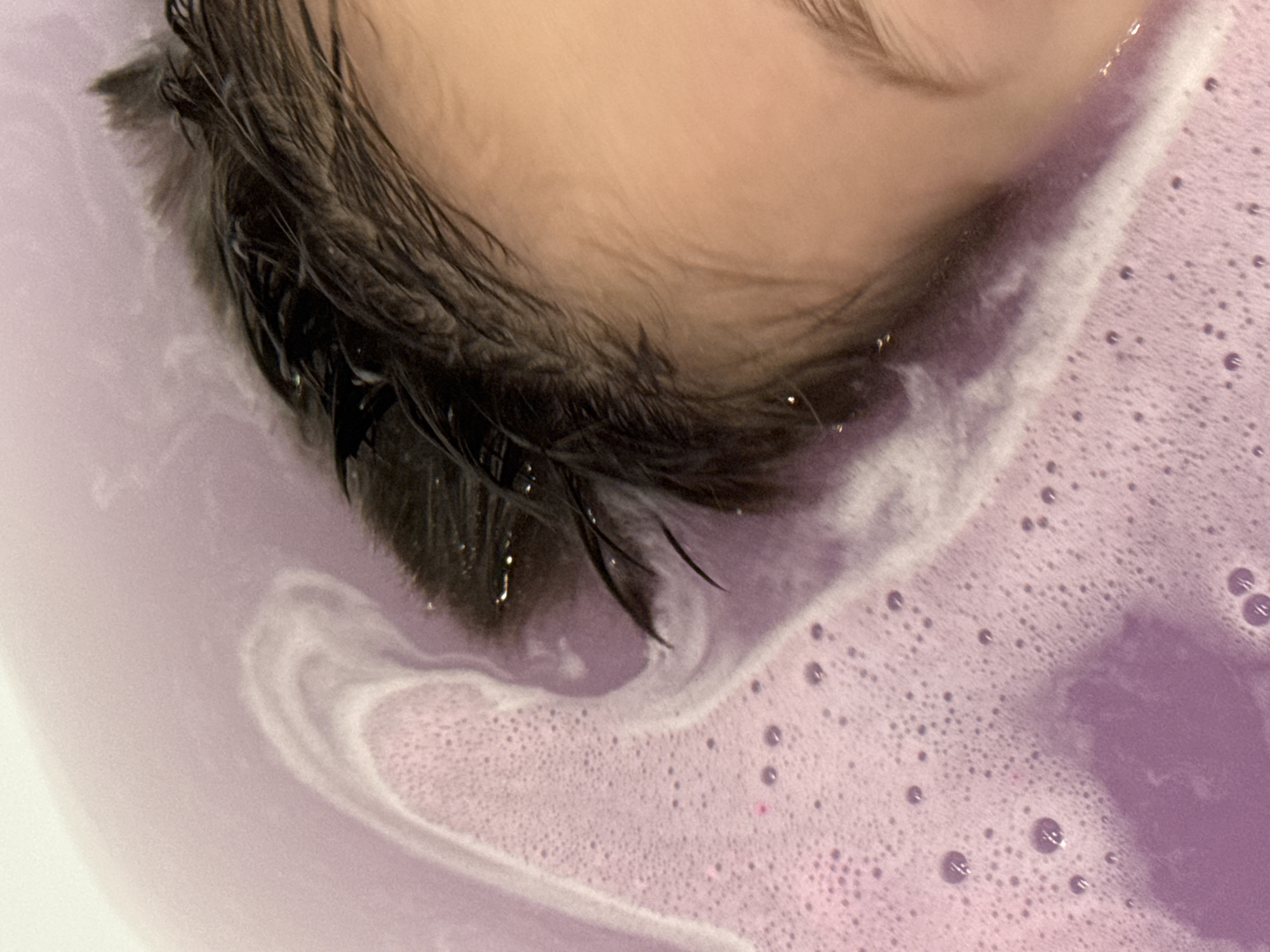 A close-up, overhead shot shows the head and hair of a person submerged in a purple, bubbly bath. The water has a light purple hue and is foamy, with small bubbles scattered across the surface. Swirls of white foam create a textured pattern in the water. The person's dark, wet hair is visible, slicked back and glistening. A portion of their forehead and eye are also visible, with smooth skin and a hint of an eyebrow. The lighting is soft, creating a relaxed atmosphere. The scene is domestic, suggesting a private bathing experience. There is no information in the image to indicate it is located in Lyubertsy, Russia.