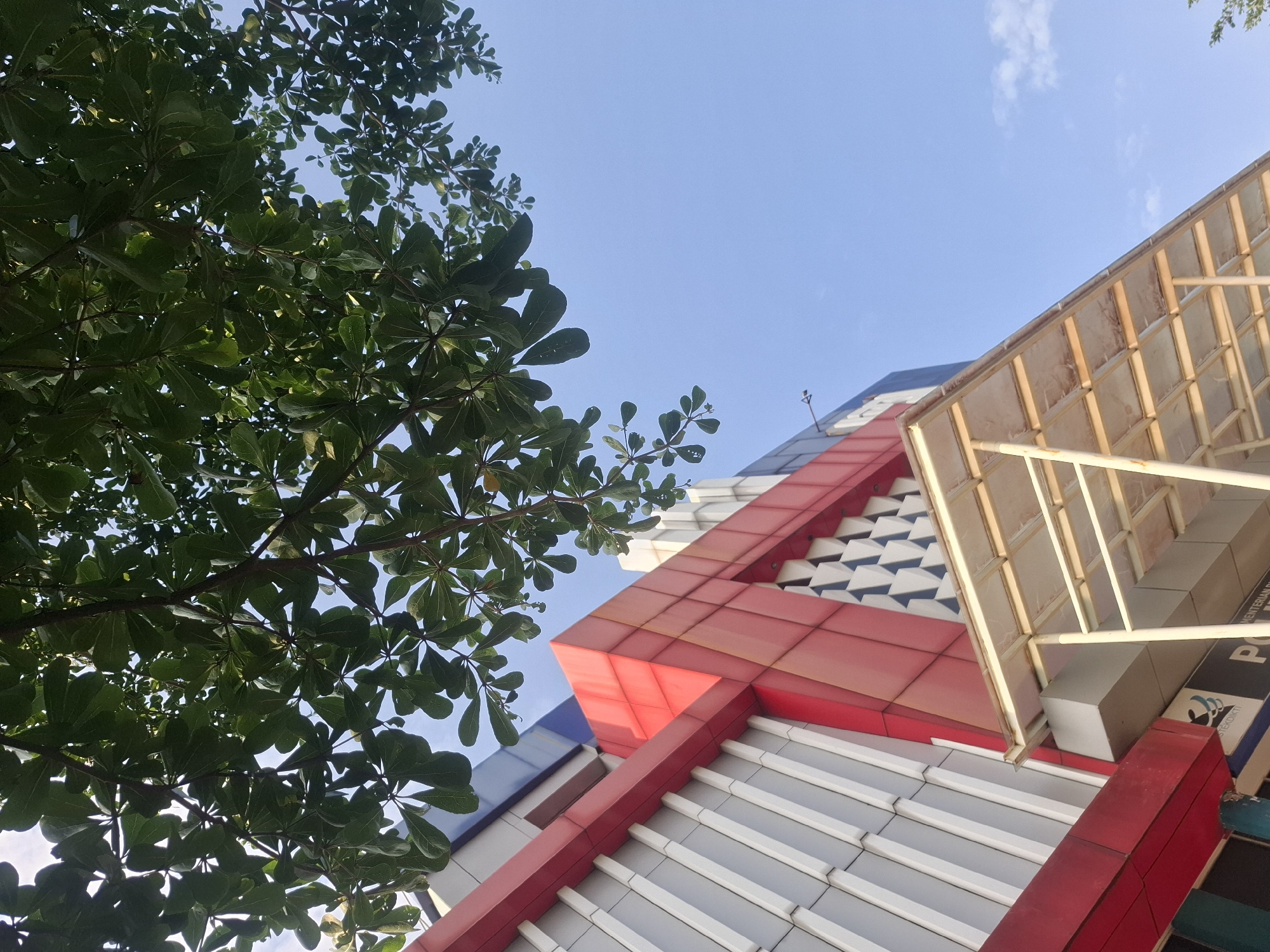 A low-angle, full-shot photograph captures the exterior of a modern building in Medan, Indonesia, under a clear blue sky. The building's facade is a striking composition of geometric shapes and colors, primarily featuring a combination of gray and white vertical slats, accented by bold red geometric panels. A white, open-sided canopy with a corrugated roof extends from the building's entrance area. Overhead, the lush green foliage of a tree branches across the top of the frame, its leaves partially obscuring the sky. The scene appears to be an outdoor public space, with the building's architectural style suggesting it could be a government office or a commercial establishment. The lighting suggests it is daytime, likely late afternoon due to the warm, soft light illuminating the canopy. No people are visible in the frame.