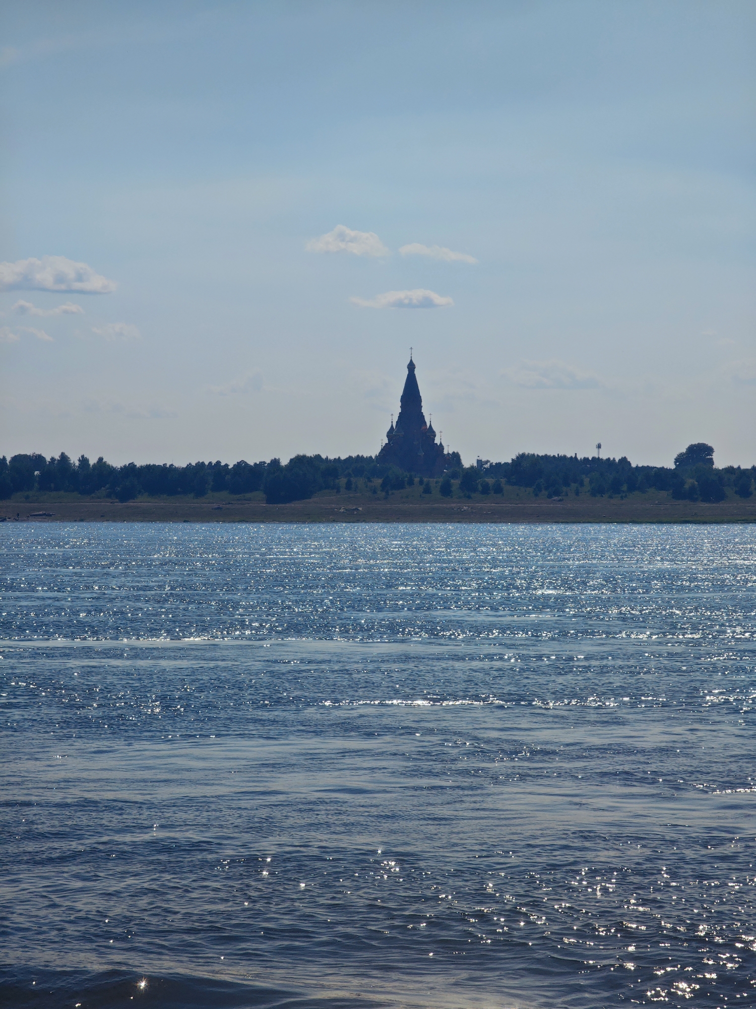 A body of water, likely a river, dominates the foreground of this image, its surface shimmering with sunlight. In the middle ground, a dense line of trees forms a dark silhouette against a hazy sky. Perched atop this tree line, a large, dark, domed structure, possibly a church or monastery, is visible in the distance, its silhouette clearly defined against the sky. The sky itself is a pale blue, with a few scattered, wispy clouds. The overall impression is one of a tranquil, possibly remote, landscape on a sunny day. The church-like structure suggests a possible location in or near a settlement in Russia. Based on the architecture of the building visible in the image and the landscape, it is highly probable that this is the Church of the Assumption of the Virgin Mary, located in the city of Lesosibirsk, Russia.