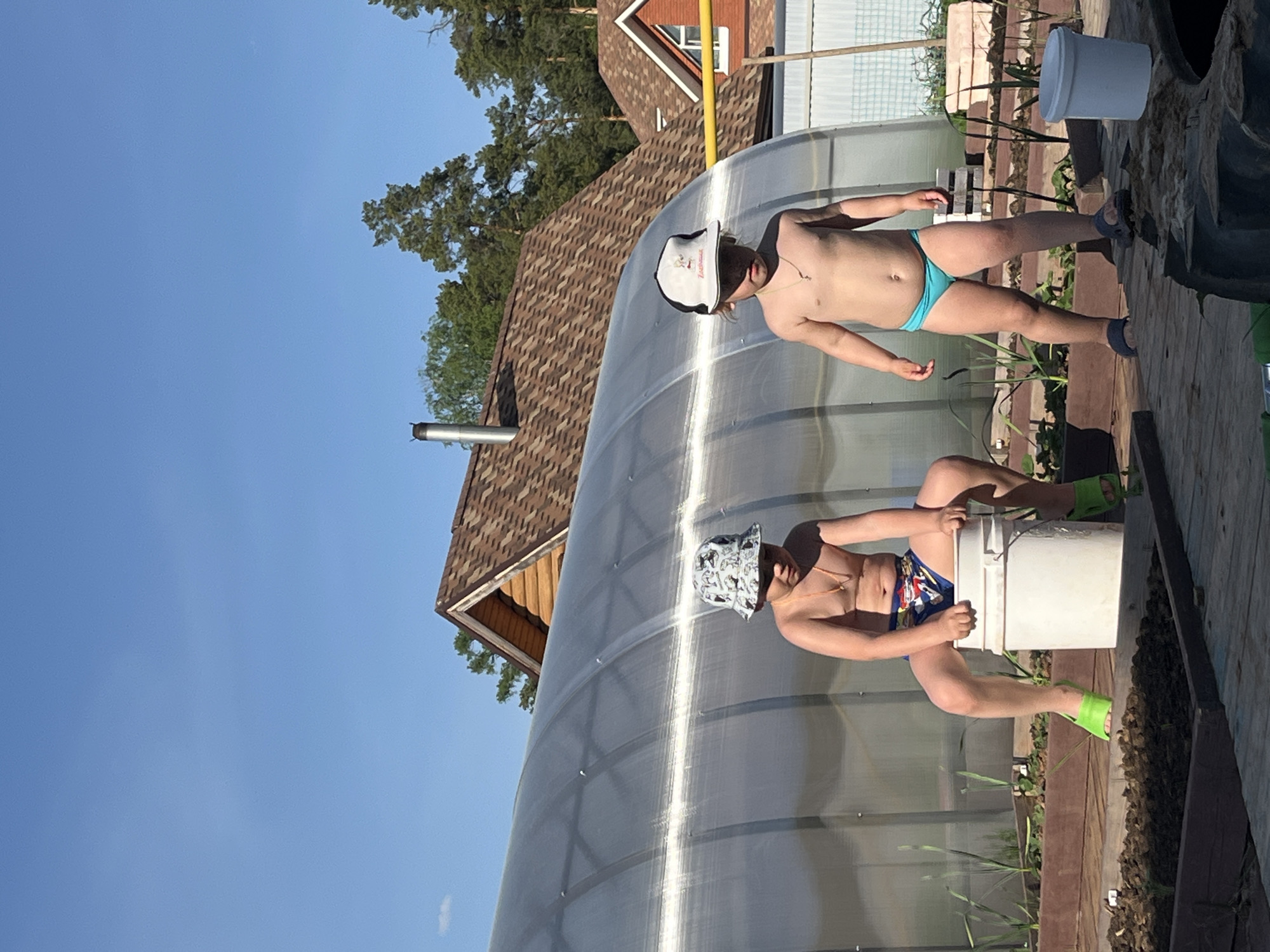 The image shows two shirtless individuals in swimwear standing outside a greenhouse. One person is wearing a white cap and blue swimwear, while the other is wearing a patterned bucket hat and patterned swimwear, standing on a bucket. The location appears to be in a rural or suburban setting, possibly Chaikovsky, Russia, based on the surroundings and context of the image. The greenhouse is made of a translucent material. The background includes a house with a brown roof, trees, and a clear blue sky.