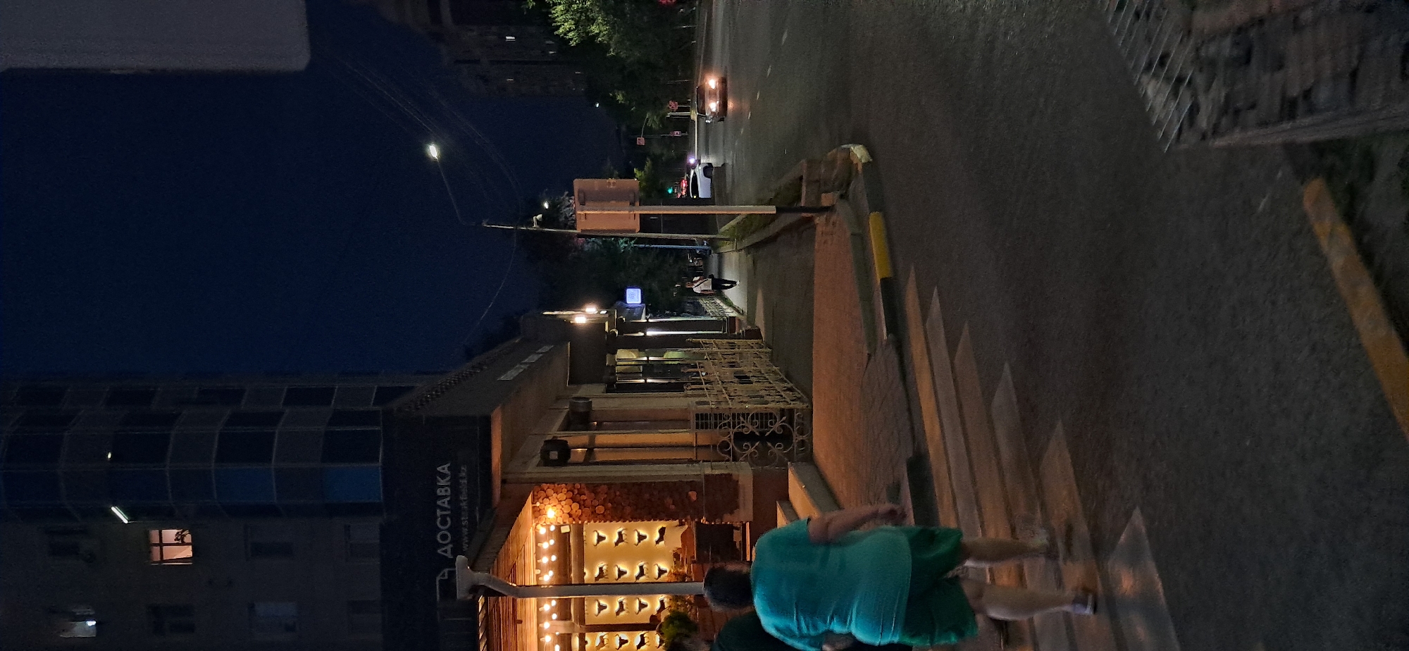 An outdoor, eye-level, vertical shot captures a dimly lit city street scene at dusk or night. On the left, a multi-story building with windows and a modern, curved facade looms in the background. In the mid-ground, a building with a sign that reads "ДОСТАВКА" (Delivery) and decorative lights is visible. A sidewalk runs alongside this building, and a person in a dark shirt and light pants is walking away from the camera. A crosswalk is in the foreground, with a person wearing a teal shirt and green shorts walking across it. The street to the right has a few cars, one with its headlights on. Trees line the right side of the street, and the sky is a deep blue.