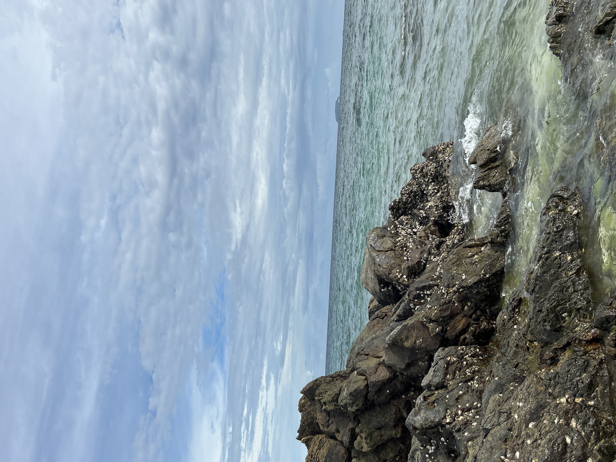 This media file captures a scenic coastal view in Tambon Phru Nai, Thailand. The image is oriented vertically, presenting a dynamic split between the sky and the sea. The left side of the frame is dominated by a dramatic sky filled with layered clouds. These clouds range from soft, wispy formations to thicker, more textured bands, suggesting a day with variable weather. The hues of the sky are predominantly blues and whites, with hints of grey indicating the cloud cover.
The right side of the image depicts the ocean and a rocky shoreline. The water is a pale, translucent green, with gentle waves lapping against dark, rugged rocks. These rocks are covered in barnacles and other marine life, adding a textured and natural element to the scene. The water appears calm, with small whitecaps indicating the movement of the waves. In the distance, a faint outline of land or an island can be seen on the horizon, partially obscured by the haze.
The overall mood is one of natural beauty and tranquility. The light suggests it could be daytime, possibly late morning or early afternoon, given the brightness but also the cloud cover. There are no people or man-made objects explicitly visible, emphasizing the raw, natural environment. The scene conveys a sense of peacefulness and the vastness of nature. No text is visible in the image.