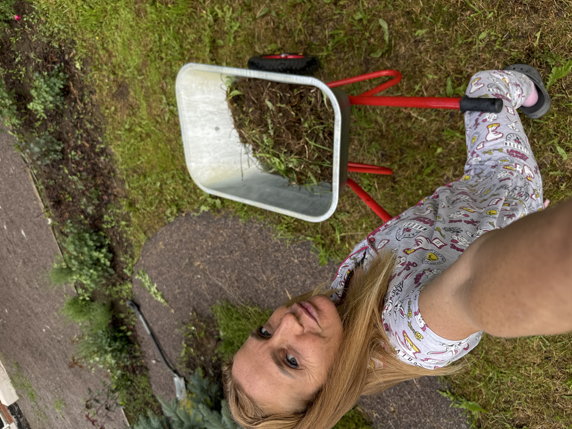 A woman is taking a selfie in a yard with a wheelbarrow. The woman has blonde hair and is wearing pajamas with cartoon characters on them. She is also wearing black clogs. The wheelbarrow is red and silver and is filled with grass clippings. The yard is green with grass and dirt. The image appears to be taken on a cloudy day. There is no visible text in the image.