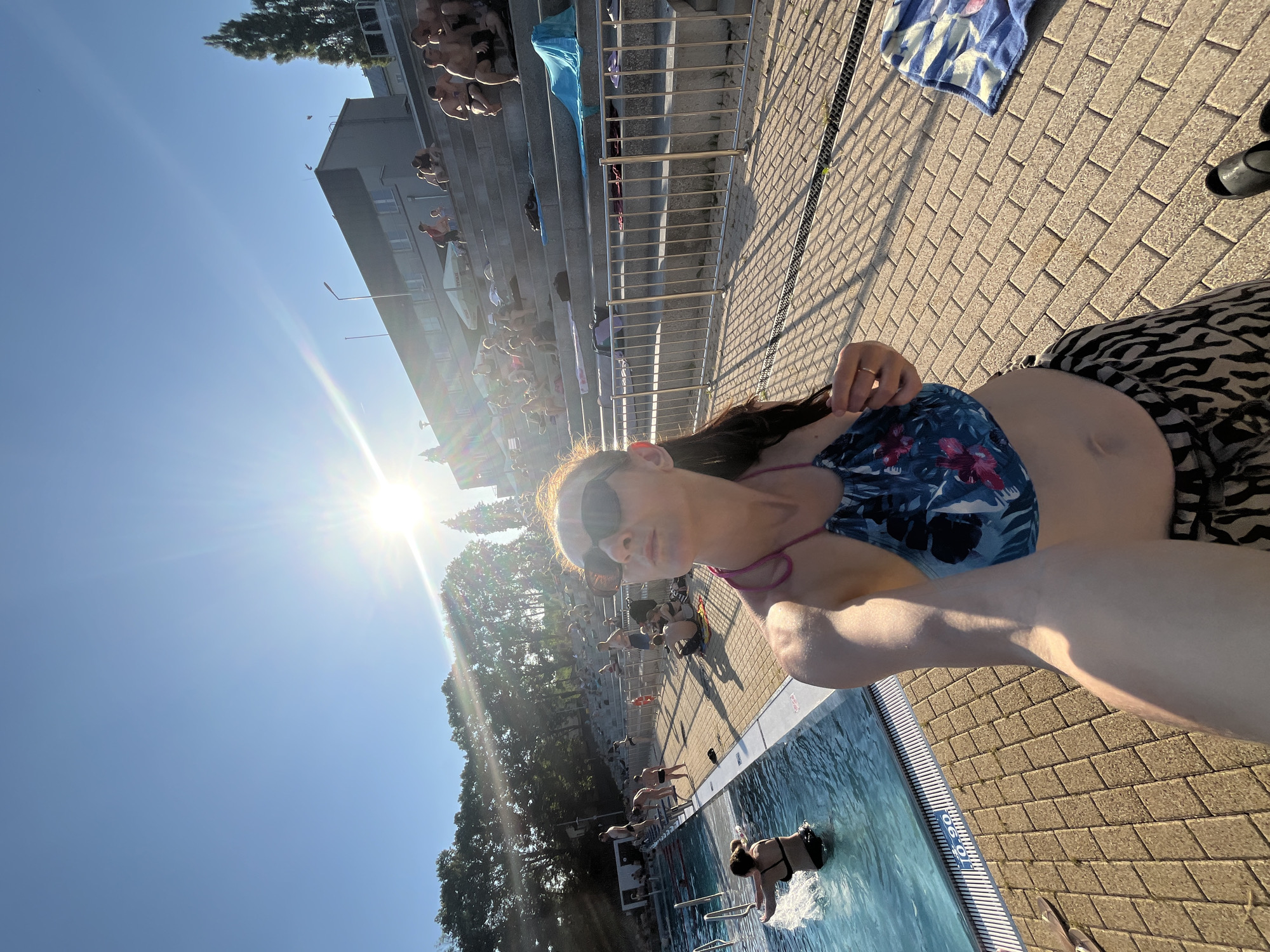 The image depicts an outdoor swimming pool area on a sunny day. In the foreground, a woman is taking a selfie. She is wearing a floral-patterned bikini top and patterned bottoms, sunglasses, and a ring on her left hand. Her right arm is extended, holding the camera. Behind her, the swimming pool is visible with a woman in the water splashing. The pool area is paved with light-colored bricks, and there are metal railings. To the right of the pool, there are tiered concrete steps filled with people sitting and sunbathing. These steps lead up to a building. Trees line the background, and the sky is clear and blue with the sun shining brightly, causing lens flare. Several towels are placed on the steps and near the pool. The environment appears to be a public recreational area.