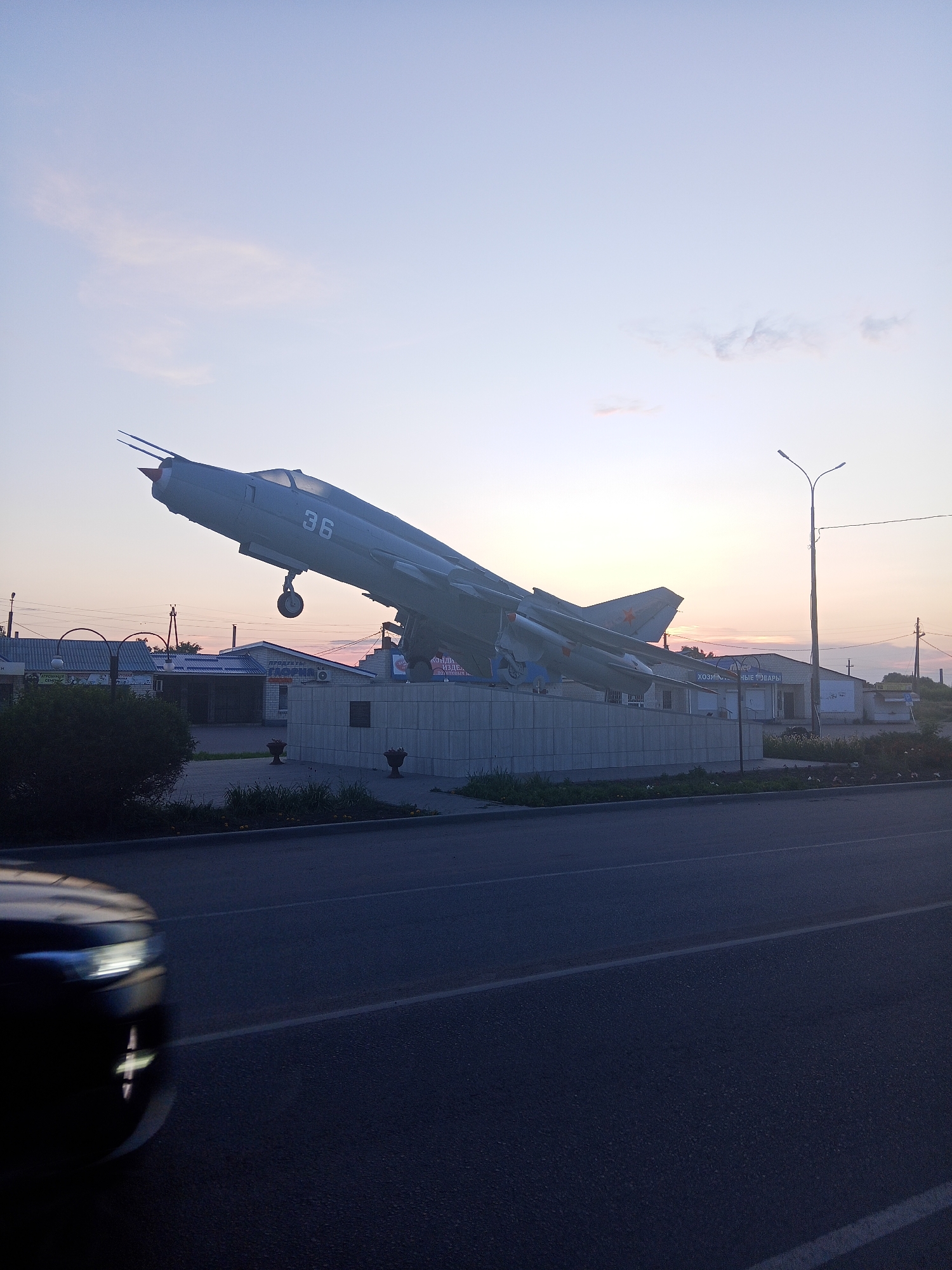 An outdoor shot captures a monument of a grey jet fighter, possibly a MiG-21, with the number 36 visible on its fuselage. The jet is mounted on a concrete pedestal in front of a row of low-rise buildings, suggesting an urban or town setting. A streetlight stands to the right of the jet. In the foreground, a dark car is blurred due to motion, driving on an asphalt road. The sky is a pale pink and blue, indicating either dawn or dusk. The location is identified as Panino, Russia.