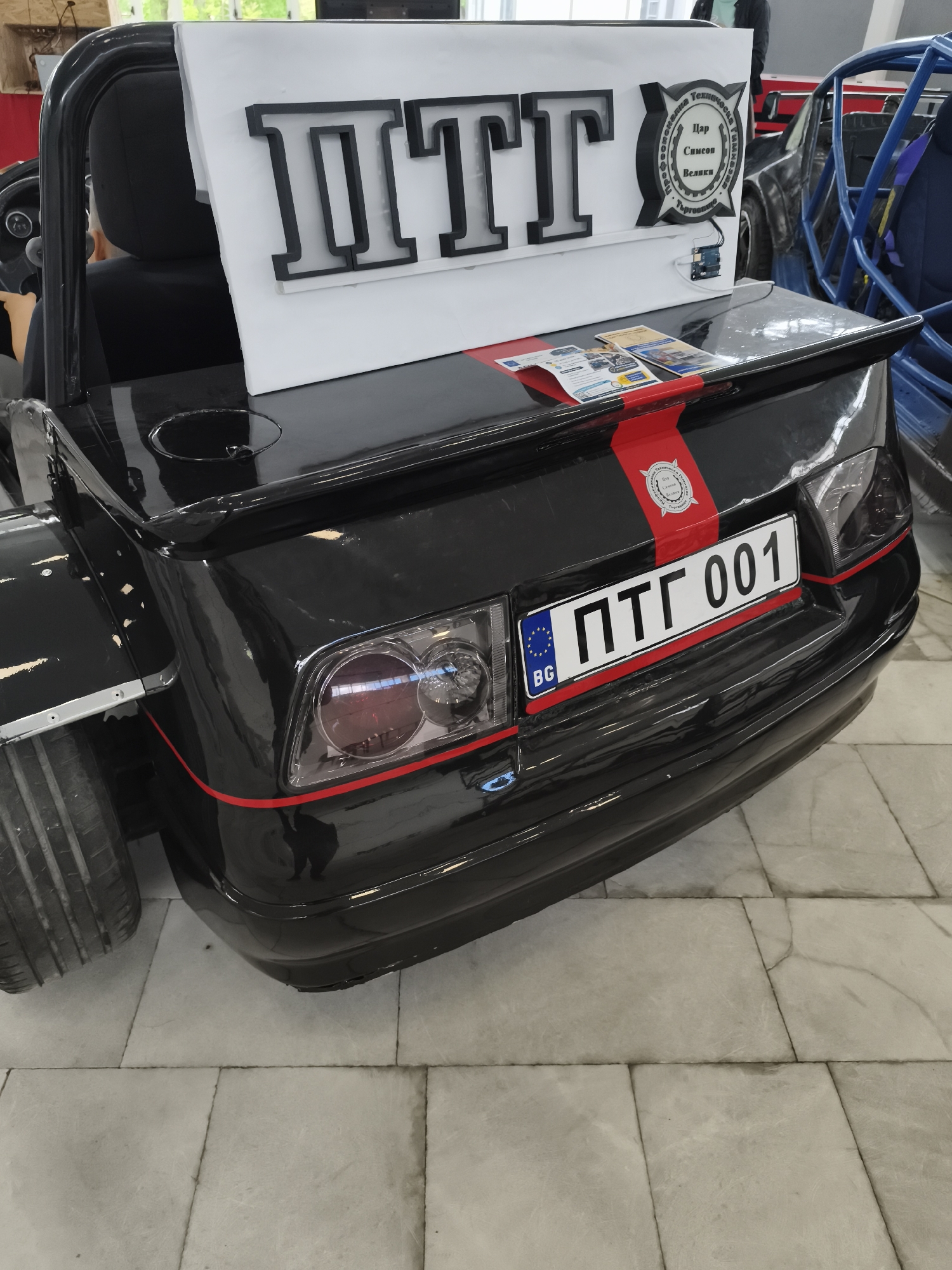The image shows the rear of a black vehicle parked indoors on a tiled floor. The vehicle has a license plate that reads "ПТГ 001" and the code "BG" indicating Bulgaria. A sign with large black letters "ITTI" is placed on the vehicle. The sign also has a logo of "Technical College Tsar Simeon Veliki, Targovishte." There is a red stripe going across the back of the vehicle. Part of another vehicle is visible on the right.