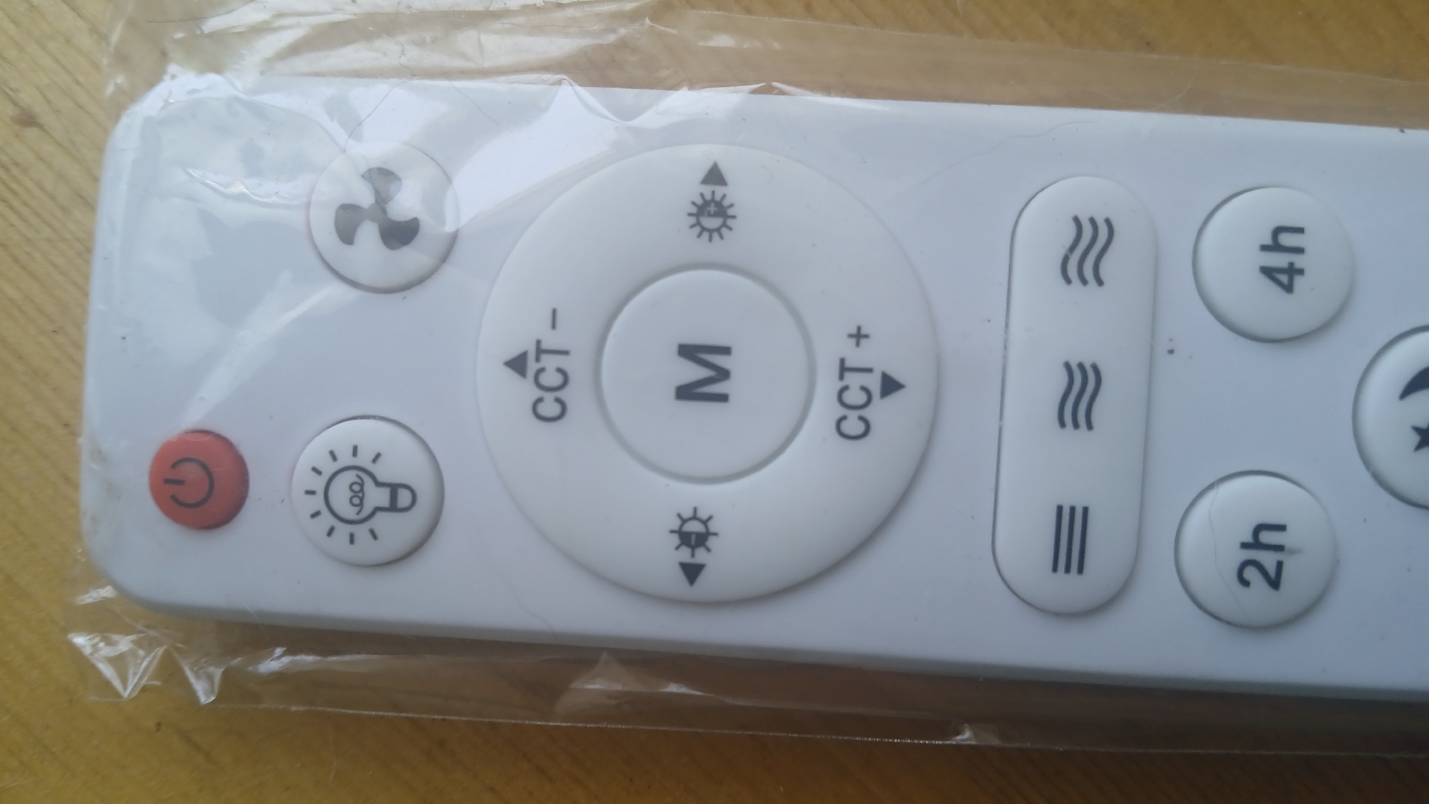 The image shows a white remote control for a light fixture, viewed from a top-down perspective. The remote is encased in clear plastic. On the remote, there are several buttons with icons and text. At the top, there is a red power button with a circular arrow. Below that, there are two circular buttons, one with a light bulb icon and another with a fan icon. In the center, there is a large circular dial with "M" in the middle, surrounded by buttons labeled "CCT -" and "CCT +" and two buttons with sun-like icons. Below the central dial are three elongated buttons with horizontal lines or wavy lines. At the bottom, there are two circular buttons labeled "2h" and "4h", and a button with a star and moon icon. The remote is lying on a wooden surface. The location of this image is unknown.