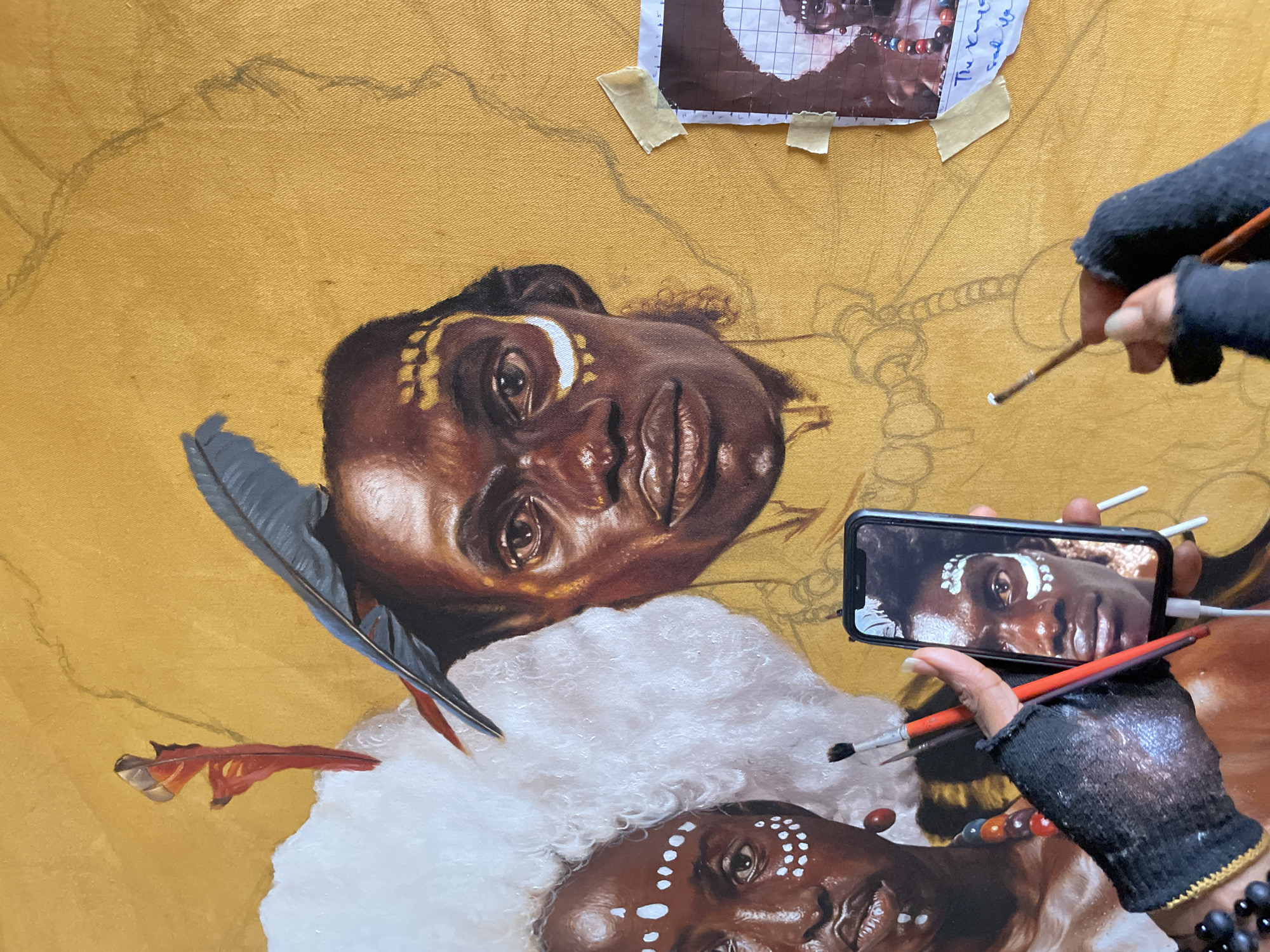 The image shows an artist painting a portrait of a person of color. The artist wears fingerless gloves and holds a phone displaying a reference image of the subject they are painting. The painting is in progress on a gold-colored canvas. The subject has decorative markings on their face and is adorned with a white headdress and feathers. A grid-marked reference image of the subject is taped to the canvas above the painted portrait. The location is not determinable from the image alone.