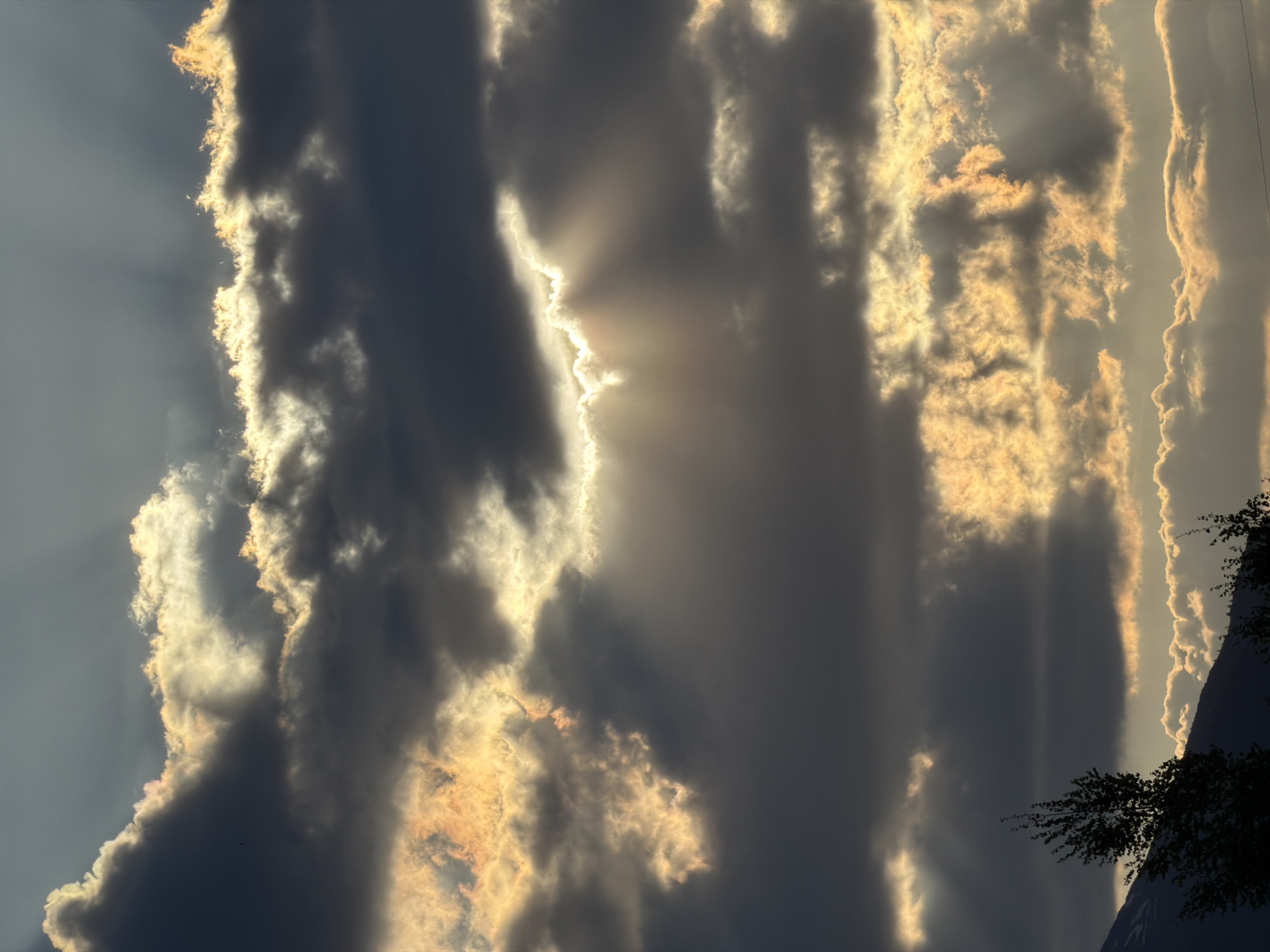 The image shows a sky filled with dark, textured clouds. Sunlight filters through the gaps, creating radiant beams. The clouds are edged with a golden light. At the bottom right corner, the silhouette of trees suggests a nearby mountain or hillside. The location is Lauševići, Montenegro.