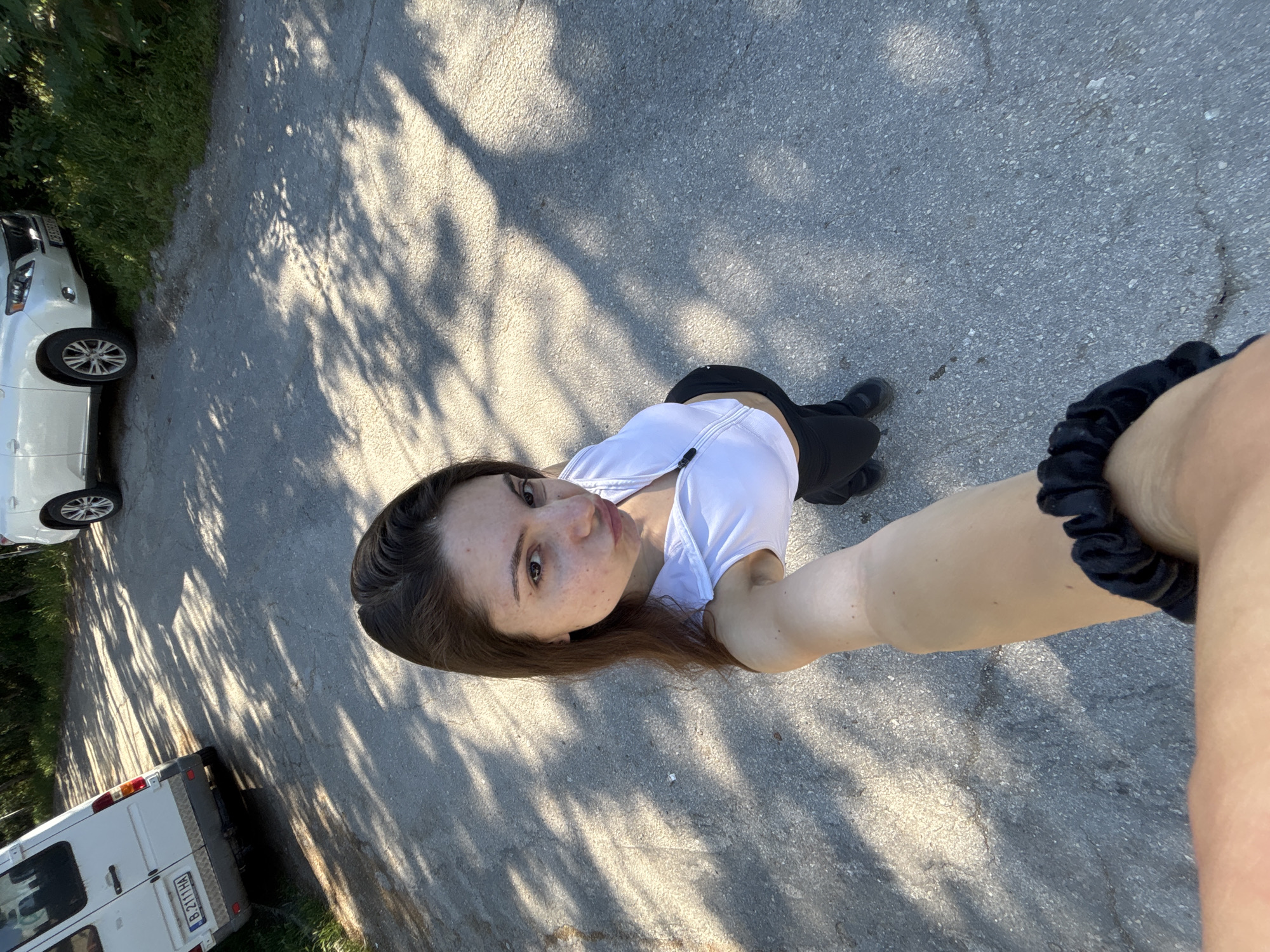 The image depicts a young woman taking a selfie on a paved road, likely in an outdoor, possibly rural or suburban, setting near Varna, Bulgaria. She is wearing a white crop top and black leggings. Her facial expression is a pout. In the background, two vehicles are visible: a white van on the left and a white SUV on the right. The road is asphalt, cracked in places, and dappled with shadows from trees. The presence of vehicles and a paved road suggests a location accessible by car, possibly on the outskirts of a town or in a park area.