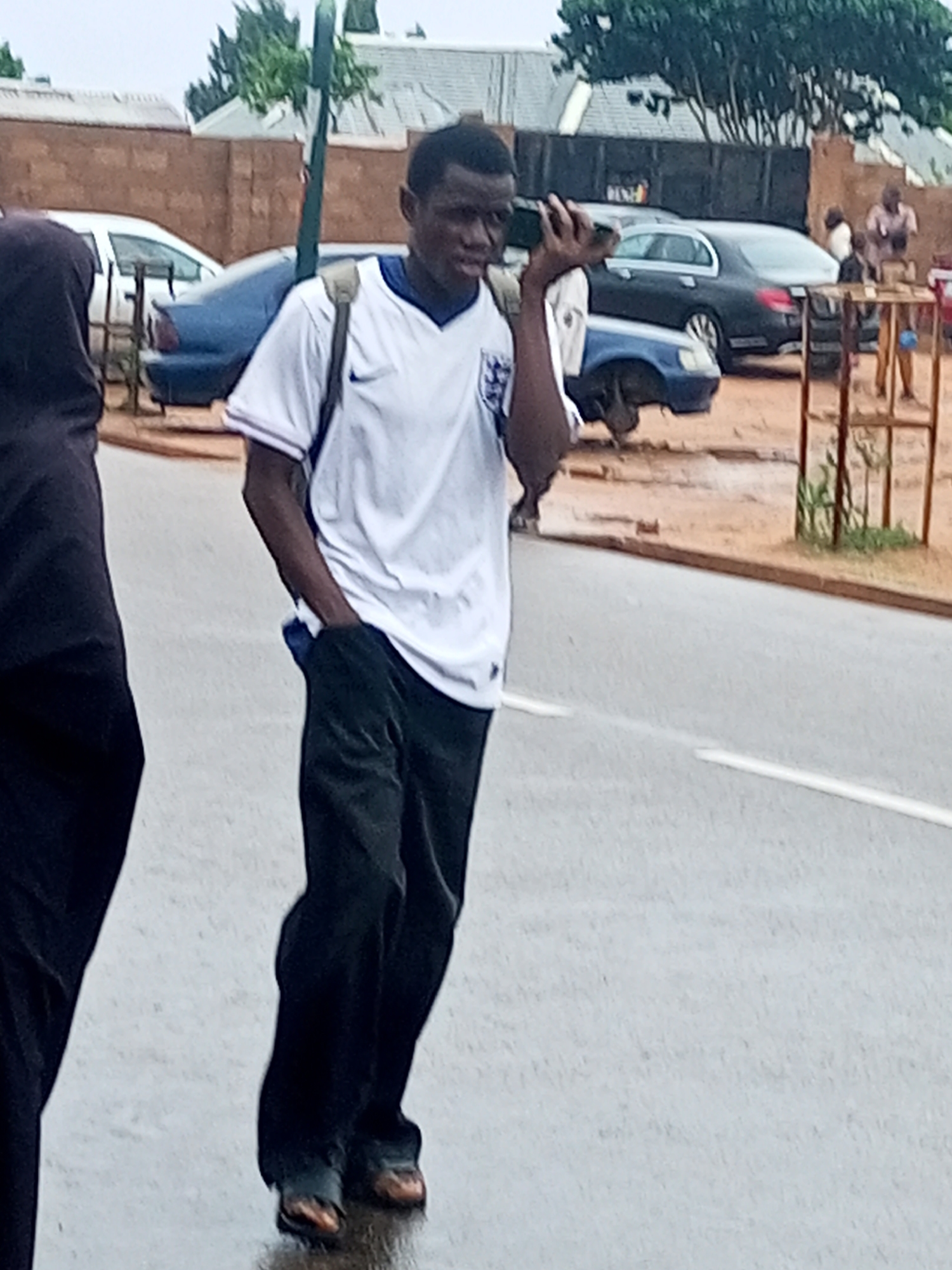 A street scene shows a young Black man walking or standing on a wet, gray asphalt road. He is in the foreground, slightly to the right of the center, wearing a white short-sleeved t-shirt with the England national football team's "Three Lions" crest on the chest, dark trousers, and brown shoes or sandals. He has a light-colored backpack slung over his shoulders. His right hand holds a dark-colored phone to his ear, suggesting he is on a call, and his left hand is in his pocket. His expression is neutral.
To the far left, a person dressed in dark clothing is partially visible, with only their torso and a leg in the frame. The road surface has a single white line. In the mid-ground, several vehicles are present: a light blue sedan is partially visible behind the main subject, and further back, a black sedan (possibly a Mercedes) is parked near a brown brick wall. Another white car is faintly seen on the far left.
The background depicts a residential or semi-urban area with various buildings, some featuring corrugated roofs, and lush green trees. A green metal pole stands upright behind the main subject. On the right side of the road, a metal frame structure, possibly part of a stall or fence, is visible, along with some blurred figures near the black car and wall. The wet appearance of the road indicates recent rain or an overcast day. Aside from the football crest, no other text is clearly legible.
