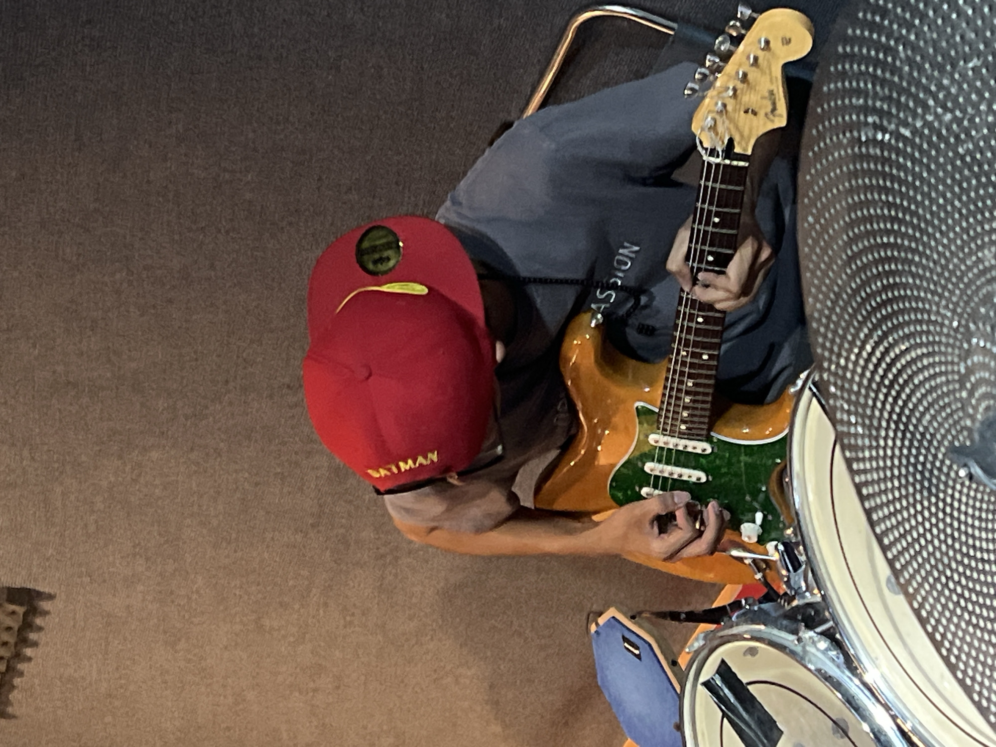 A person wearing a red "BATMAN" baseball cap and a grey t-shirt is playing a guitar. The guitar has a sunburst finish and a green pickguard. The person is seated in front of a drum set, with a snare drum and cymbals visible. The background is a textured, grey wall. The scene appears to be an indoor practice space or studio. The individual is likely in the city of Đà Nẵng, Vietnam.