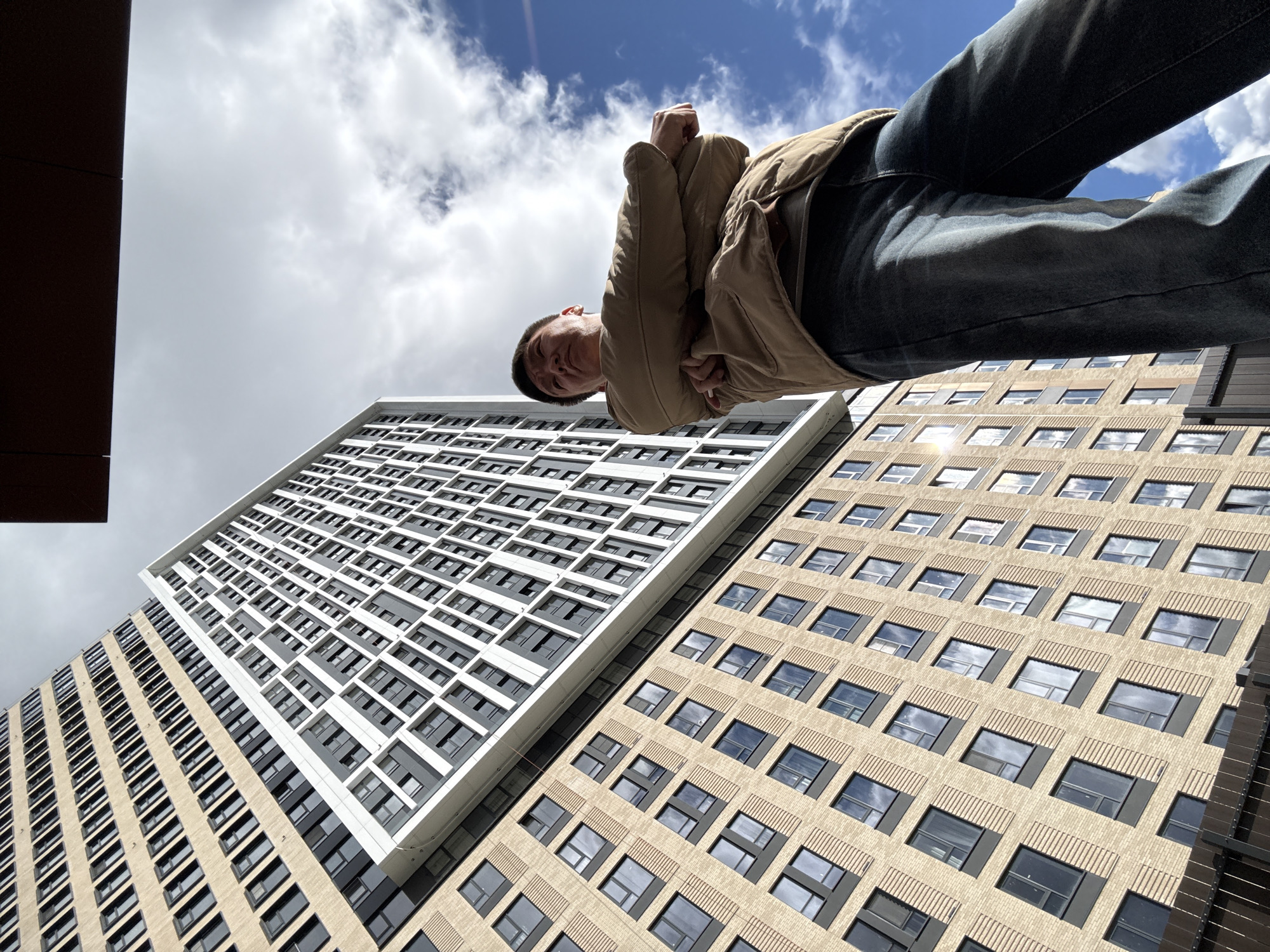 The photograph depicts a low-angle, upward-facing perspective of a person standing above and in front of two tall buildings, set against a backdrop of a cloudy blue sky. The person, likely a young adult, is wearing a light brown jacket and dark blue jeans. The angle suggests the photograph was taken from below, looking up at the subject.
The buildings are multi-story structures with a grid-like pattern of windows. One building features a light facade with a geometric design. The other building displays a darker facade with more traditional window placement. The location is Khimki, Russia.