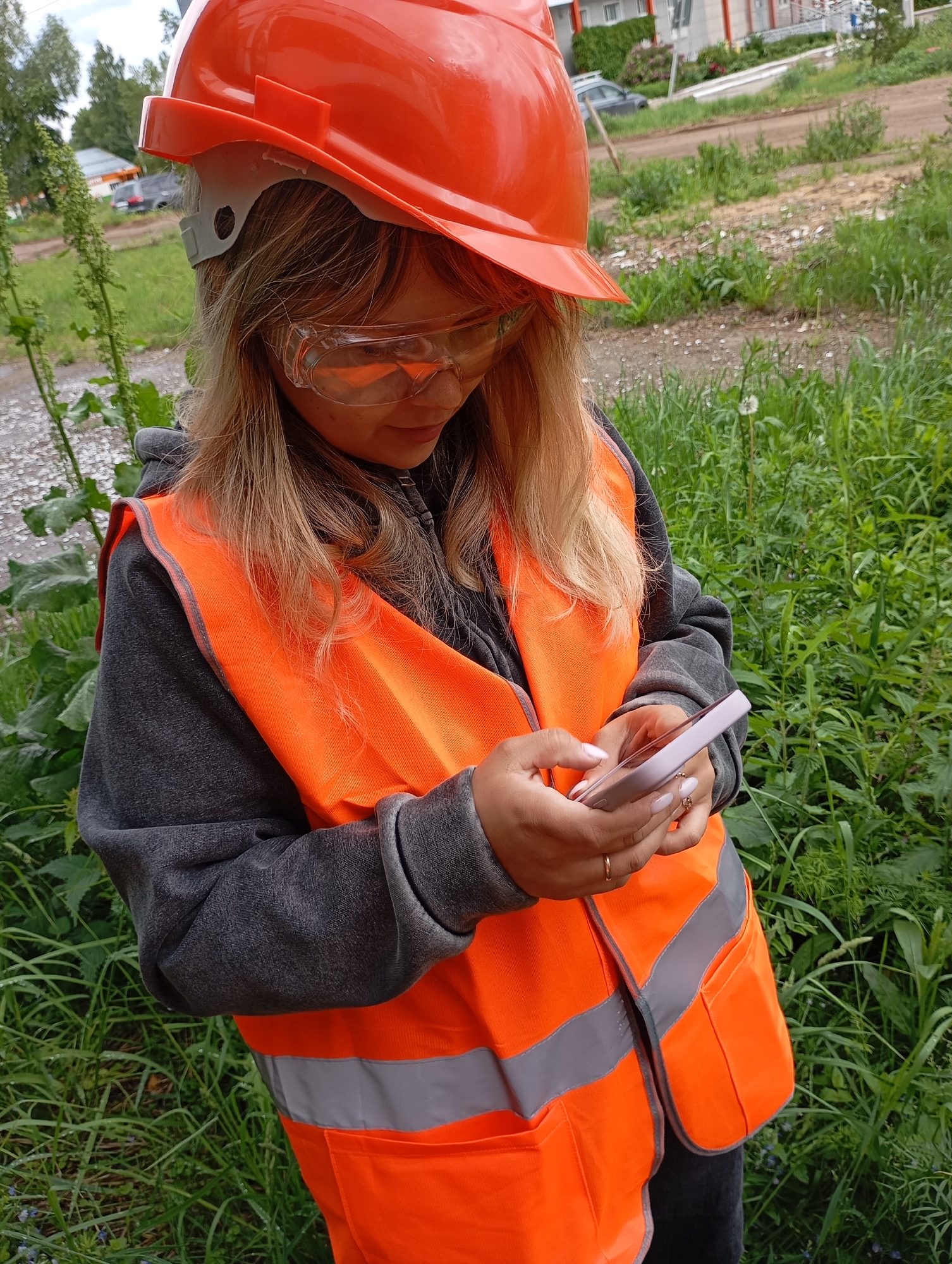 A young woman, seen from a high angle, is depicted outdoors in Chaikovsky, Russia. She wears essential safety gear, including a bright orange hard hat, clear protective goggles, and an orange high-visibility vest with reflective grey stripes over a grey hooded sweatshirt. Her blonde hair is visible beneath the hard hat.
The woman is focused on a light-colored smartphone, which she holds with both hands. She appears to be engrossed in an activity on the device, possibly checking data, communicating, or using an application related to her work. She has light-colored nail polish and a ring on her right hand.
The setting is an unpaved, grassy area, likely a field or the periphery of a development site, characterized by abundant green vegetation, tall weeds, and a dirt path or road. In the background, distant buildings or houses and a parked car are faintly visible. The scene takes place during daytime, under what appears to be an overcast or partly cloudy sky, resulting in soft, diffused natural light. Her attire suggests she is involved in construction, surveying, or fieldwork. No discernible text is visible in the image.