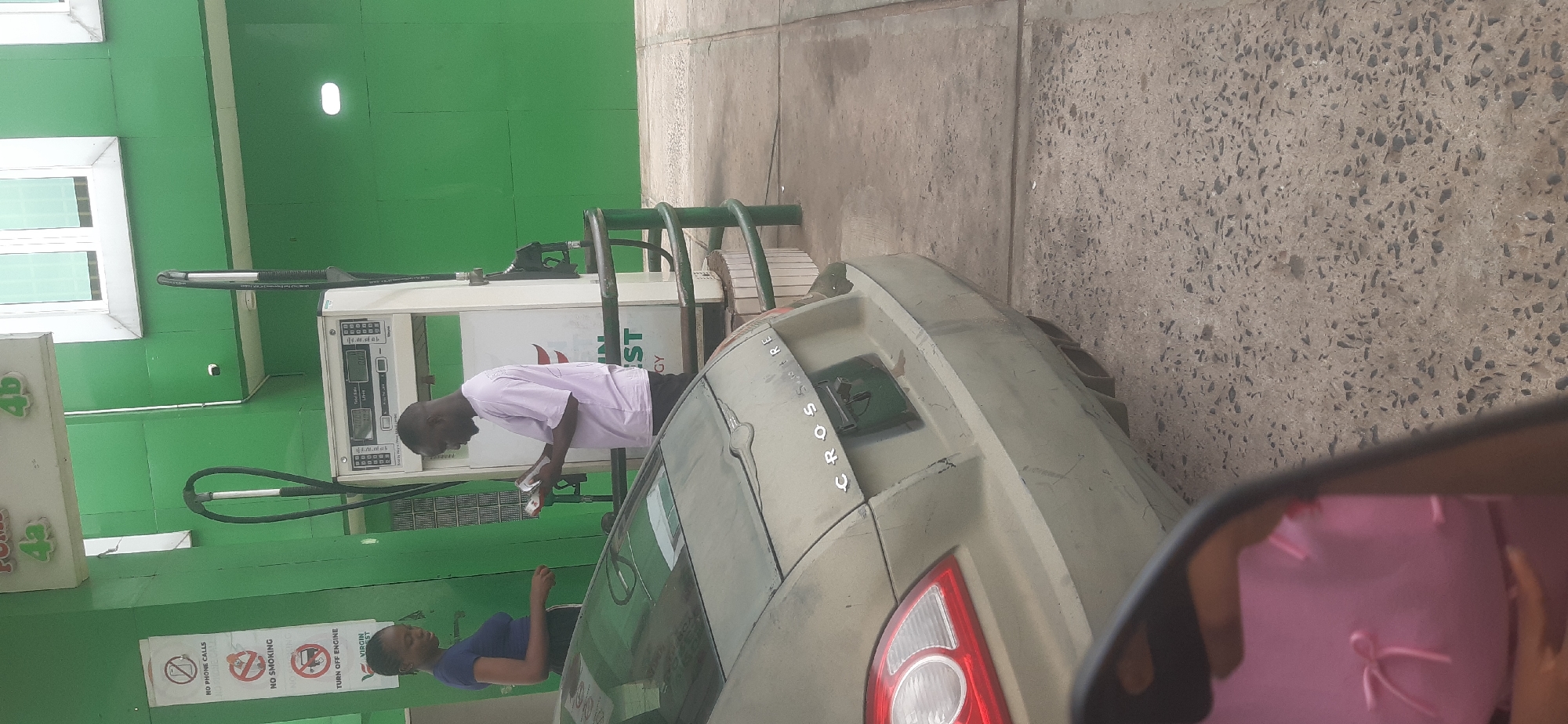 This image depicts a daytime scene at a gas station in Onitsha, Nigeria. The primary subjects are a man, a woman, and a car being refueled.
The man, dressed in a light-colored t-shirt and dark trousers, stands next to a gas pump, seemingly engaged in a transaction involving money or a card while attending to the fueling of a light-colored car. The car's rear features the text "CROSFIRE," likely referring to a "Crossfire" model. Partially visible behind the man is a woman wearing a dark blue top, also present at the station.
The setting is characterized by a bright green building or canopy structure. A gas pump is clearly visible, displaying "Total Rate" and "Litres." Several safety warnings are posted on a white panel against the green wall, including "NO PHONE CALLS," "NO SMOKING," and "TURN OFF ENGINE." Below these, a logo with the words "VIRGIN ST" (likely part of "VIRGIN ENERGY" or "VIRGIN FOREST") is visible. Additional signs on the building feature "4b" and "43."
The ground is composed of textured concrete. The photograph appears to be taken from inside another vehicle, with the lower right corner showing a reflection in a side mirror of a pink object, possibly a bag or piece of clothing. The overall activity portrays a routine stop for vehicle fueling.