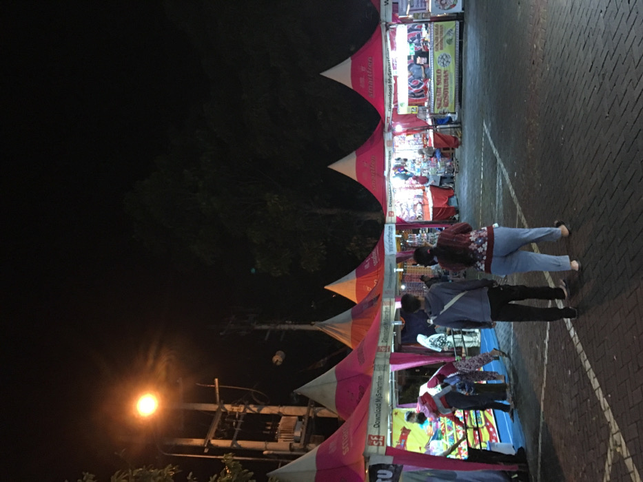 The image shows a night market in Kecamatan Batu, Indonesia, located at latitude -7.867 and longitude 112.514. The scene is illuminated by streetlights. There are five tents lined up in a row, each with a white top and pink sides. The tents are selling a variety of goods. There are several people walking around and browsing the stalls. Some people are shopping and others are looking at the goods. There are two men and two women. One of the women is wearing a long-sleeved shirt and pants. The other woman is wearing a black long-sleeved shirt and dark jeans. One of the men is wearing a blue shirt and jeans, while the other is wearing a gray shirt and pants. The ground is covered in brick pavers.