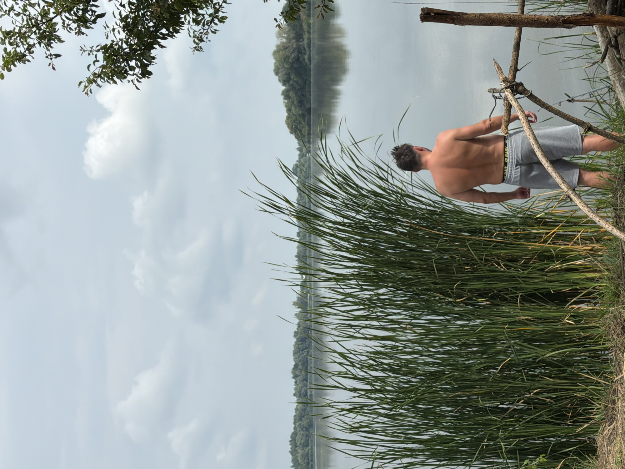 A man with short brown hair and a muscular build stands facing away from the camera, looking out at a calm body of water. He is wearing gray athletic shorts and has his back to the viewer. The man is positioned to the right of a dense stand of tall, green reeds that dominate the foreground. Behind the reeds, the water stretches out, reflecting a cloudy sky. The far bank of the water is lined with trees, appearing as a dark green band against the horizon. The overall environment suggests a natural, outdoor setting, possibly a lake or riverbank in Ruda, Ukraine. The scene is captured in natural daylight under an overcast sky.