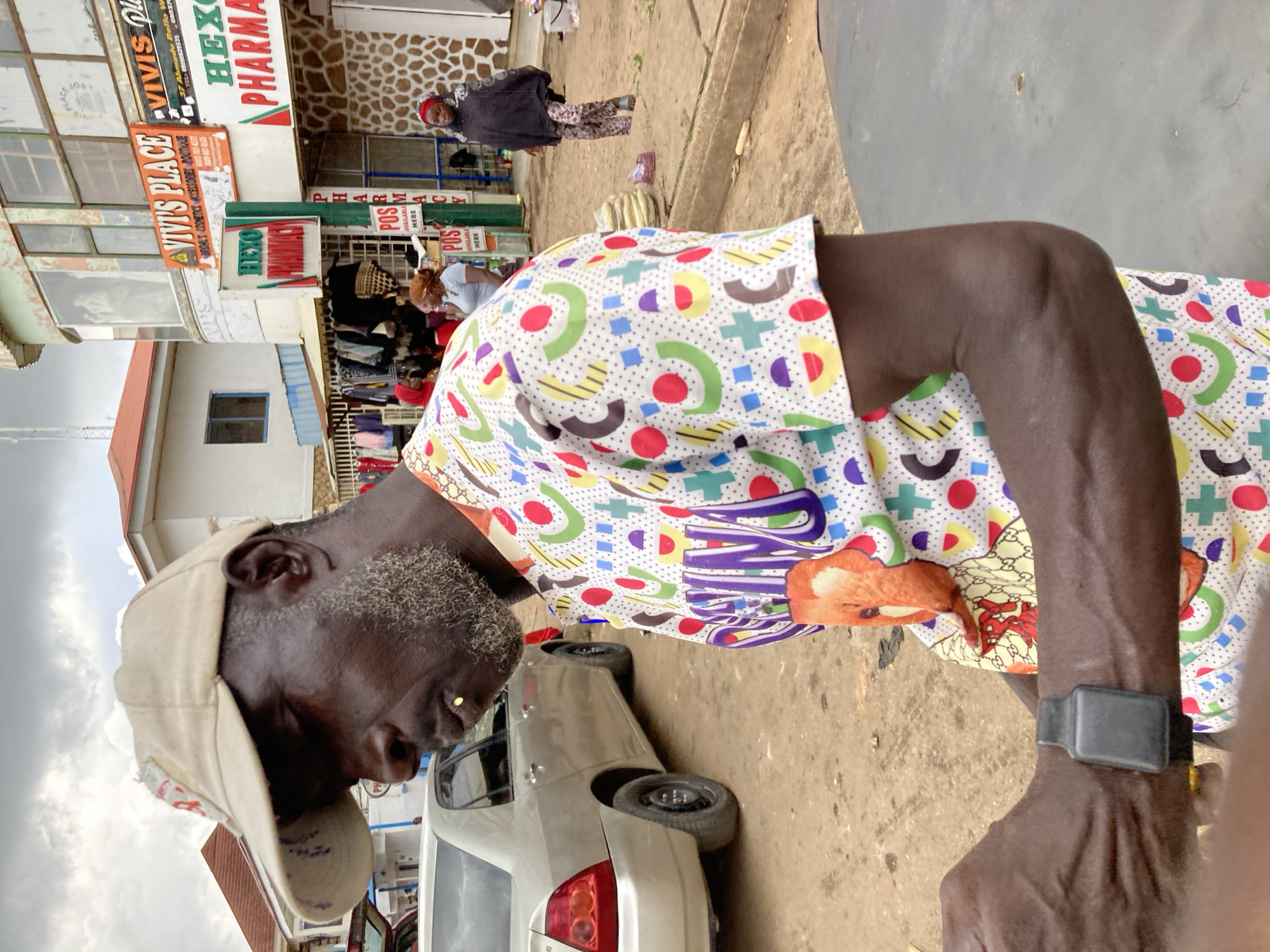The image depicts a street scene in Jos, Nigeria, with a focus on a man in the foreground. He is wearing a beige baseball cap and a colorful, patterned shirt featuring geometric shapes and cartoonish figures. His face is turned downwards, and he has grey stubble around his mouth. A silver car is visible behind him, and further in the background, several shops line the street. Signs for "VIVIS PLACE," "HEX PHARMACY," and "POS AVAILABLE HERE" are clearly visible, suggesting a commercial area. The overall lighting suggests it is daytime, possibly overcast due to the cloudy sky. The scene appears to be a typical bustling street in a Nigerian town, with people and commerce intertwined. The man's attire and the presence of various shops indicate daily life activities in this location.