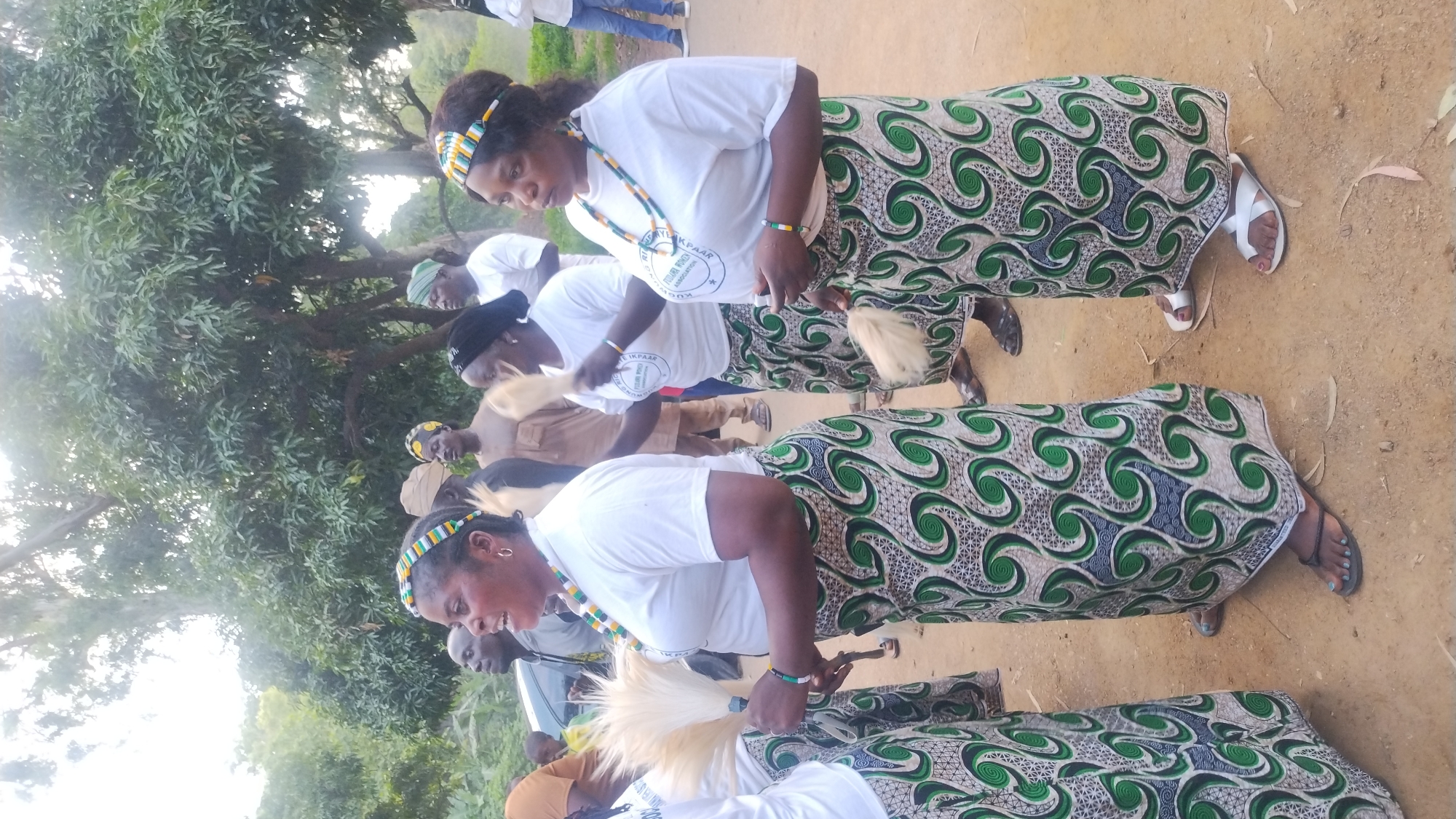 A group of women in traditional attire are standing outdoors. The women are wearing white t-shirts and long skirts with a green and white patterned fabric. Some are wearing beaded necklaces and headbands. They are holding white feathery objects. The environment appears to be a rural or semi-rural area with trees and dirt ground. The women seem to be participating in some sort of cultural event or ceremony. It is not possible to determine the specific location within Jos, Nigeria based on the image.