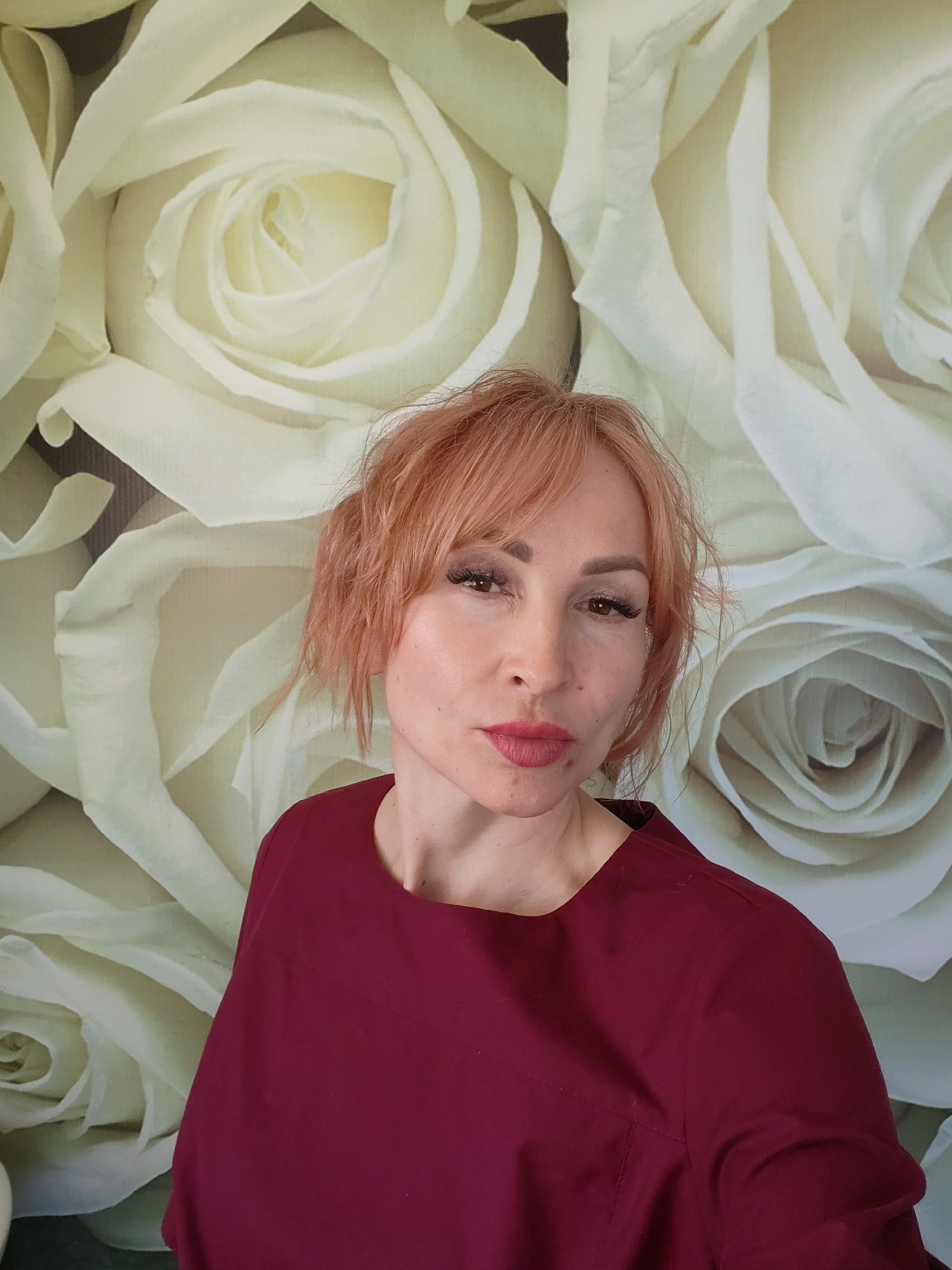 A woman with short, reddish-blonde hair poses against a backdrop of large, pale roses. She is wearing a dark red garment with a crew neck. The location is the city of Chaikovsky, Russia. The lighting is soft and even. Her expression is somewhat neutral, with a slight pursing of the lips. She appears to be facing the camera directly. Her makeup is visible, including eyeliner and lipstick.