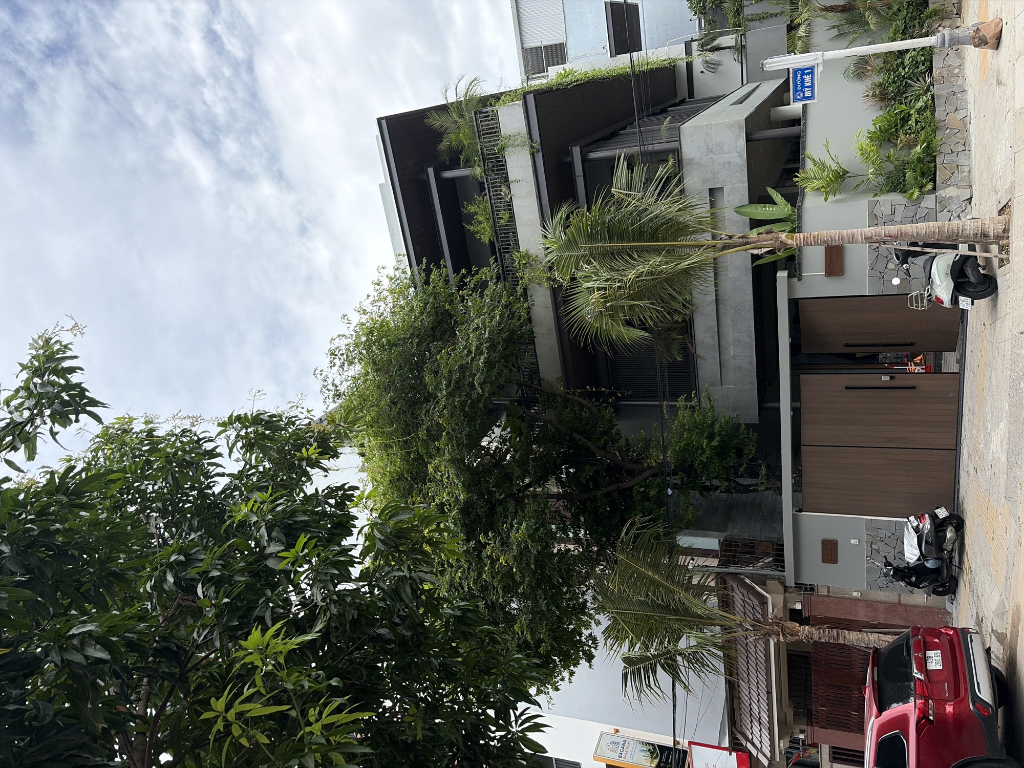The image depicts a modern, multi-story building with extensive greenery cascading from its balconies and rooftops, located on My Khe 1 street in the city of Da Nang, Vietnam. The building has a contemporary design featuring concrete, dark metal accents, and large glass windows. A large tree with lush foliage dominates the left side of the frame, obscuring parts of the building. In the foreground, a red SUV is parked to the left, and a light-colored scooter is parked in front of a large wooden gate. A street sign reading "ĐƯỜNG MY KHE 1" is visible on a lamppost. The sky is overcast with scattered clouds. The scene appears to be on a street with a sidewalk and a paved road.