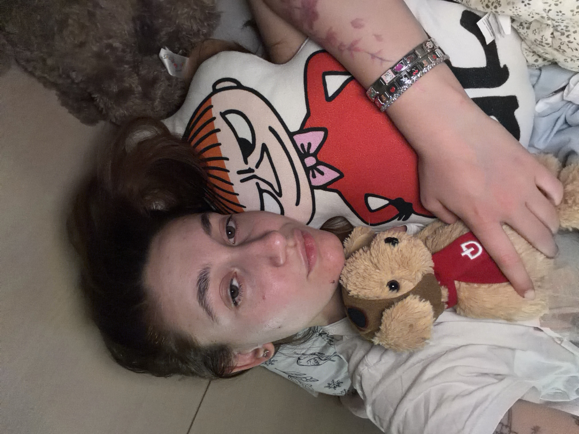 A young person, likely female, with dark brown hair, is pictured lying down on their back, looking directly at the camera. The image was taken indoors in Sofia, Bulgaria. Their expression is neutral, with a slight flush on their cheeks and an earring visible in their left ear.
The person's right arm is bent, and a decorative, sparkly bracelet with square and rectangular elements adorns their wrist. Faint red or purple marks are visible on their right forearm.
Several stuffed items surround the person. A large, fluffy, dark brown stuffed animal, possibly a bear, is partially visible in the upper left, with a white tag showing a partial "S" and other text. Underneath the person's head is a stylized plush pillow shaped like a cartoon character, featuring orange-striped hair, a prominent nose, and a red body with a pink bow. Resting near their right shoulder and under their arm is a small, light brown stuffed dog, wearing a red vest with a white letter "D" or "G" logo. The person's hand is gently placed on this small dog.
The background consists of a light-colored, possibly tiled, floor and various light-colored fabrics, including a patterned one beneath their head and a plain white one under the stuffed dog, suggesting a casual, relaxed indoor setting.