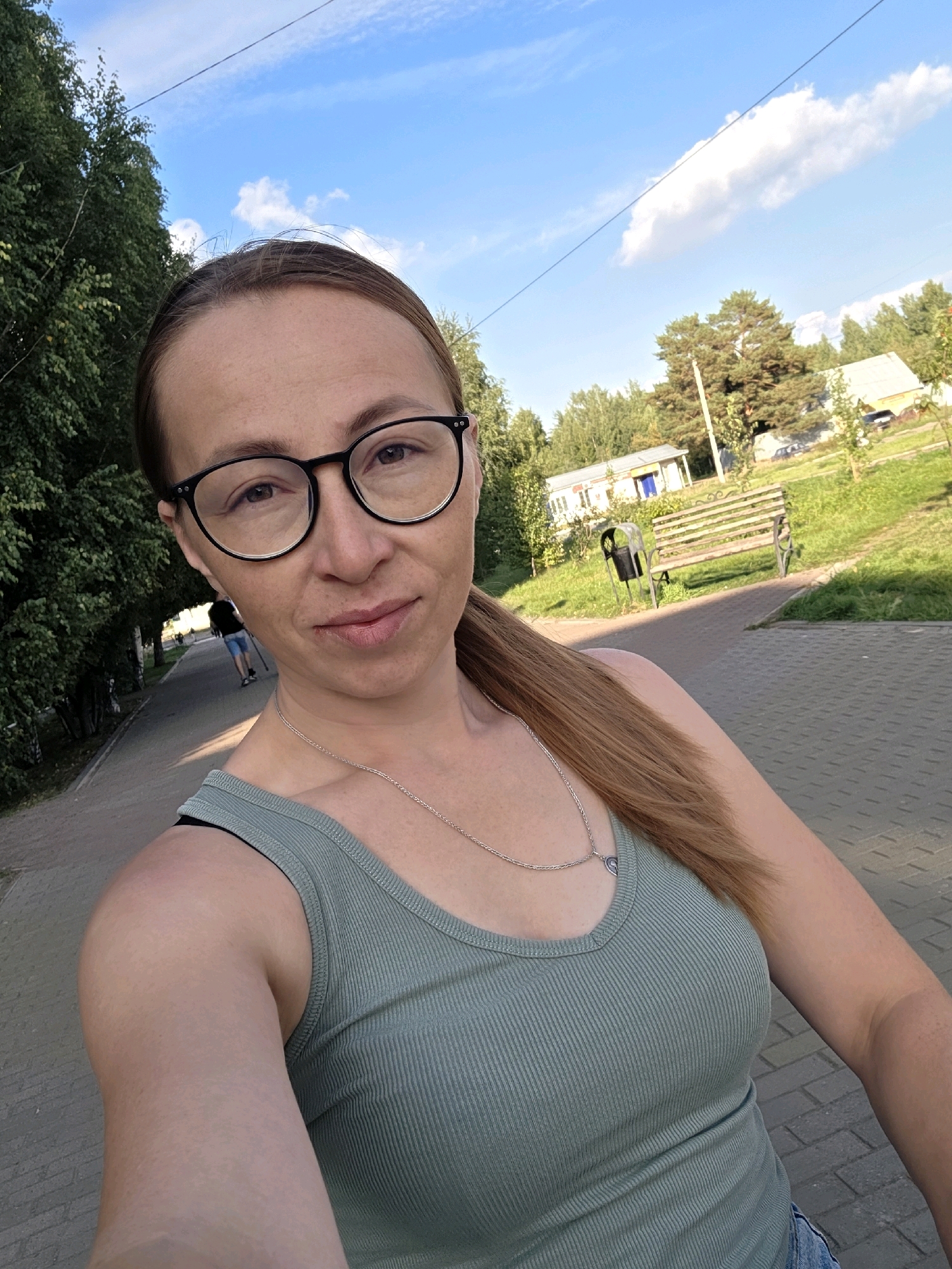 A woman is taking a selfie outdoors on a paved walkway, possibly in Chaikovsky, Russia, given the context. The woman is wearing glasses, a green tank top, and a silver necklace with a heart-shaped pendant. Her hair is long and reddish-brown, pulled back into a ponytail. In the background, there is a paved walkway lined with trees on the left and a grassy area with a park bench and a trash can on the right. Further in the background, there are some buildings and more trees under a blue sky with scattered clouds. A person is visible walking in the distance on the walkway. The lighting suggests it is daytime, possibly late afternoon, with sunlight casting shadows.