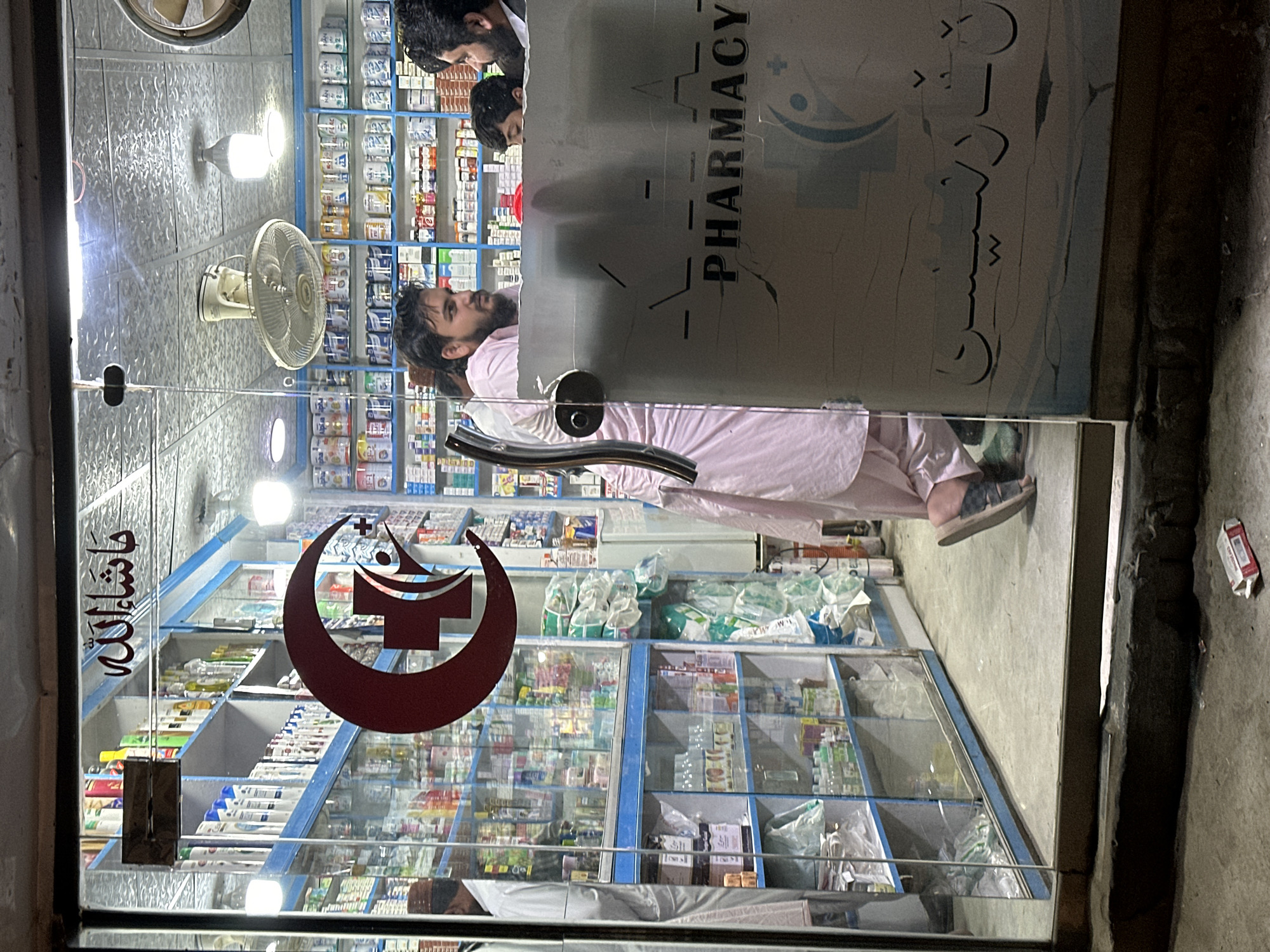The image depicts the exterior of a pharmacy located in Quetta City Tehsil, Pakistan. The pharmacy's glass door is the primary focus, displaying the word "PHARMACY" along with an emblem. Additional signage in Urdu is visible on the door. Inside the pharmacy, shelves are stocked with various medications and pharmaceutical products. A person is visible inside the pharmacy, seemingly browsing. Two additional individuals are seen near the top of the frame, peering into the pharmacy. An electric fan is mounted on the ceiling inside. The exterior environment appears to be urban, with a narrow sidewalk and a wall on the right side of the frame. A discarded cigarette box is lying on the sidewalk.