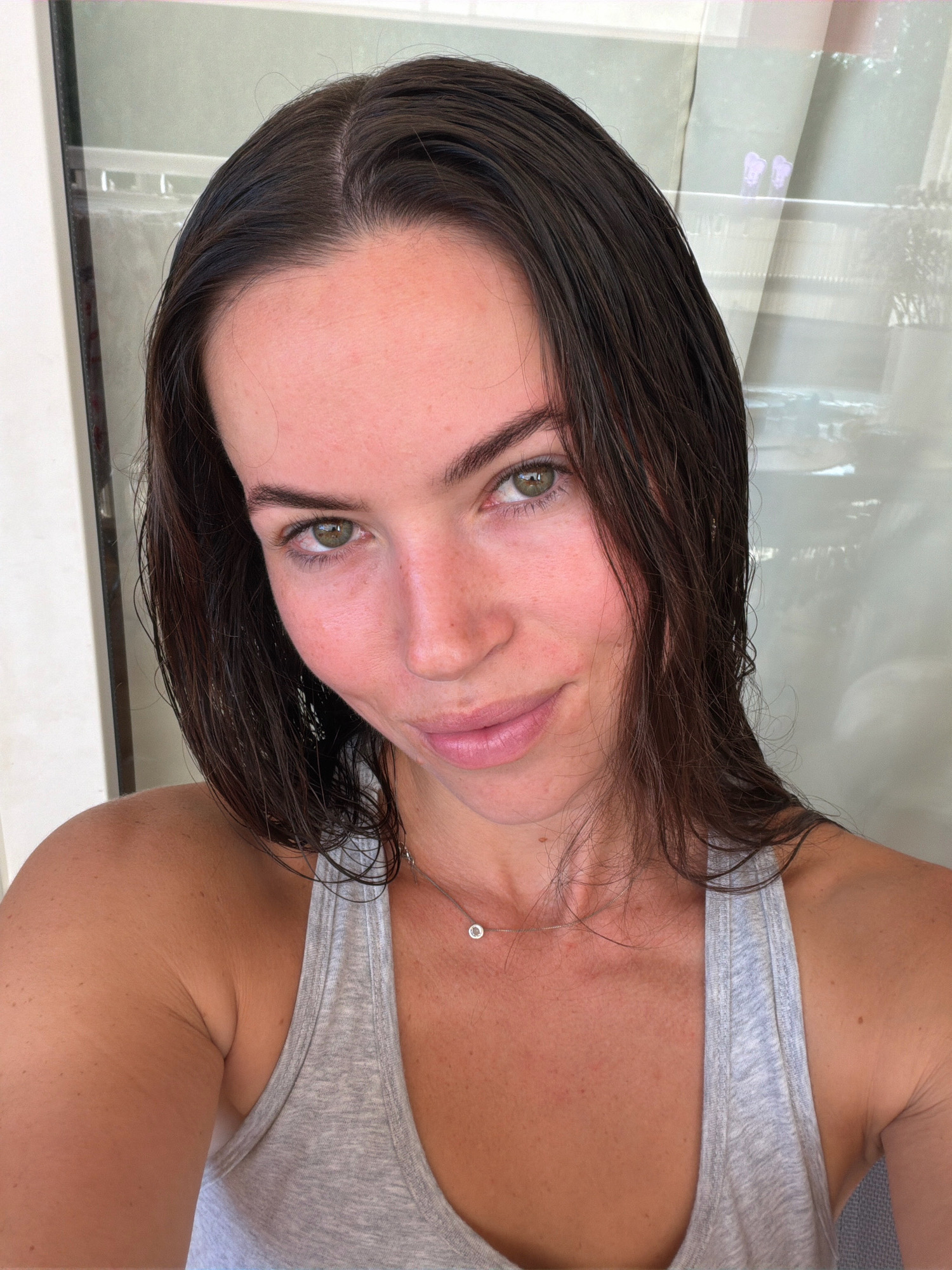 A young woman with dark, wet hair and green eyes is looking at the camera. She is wearing a grey tank top and a thin silver necklace with a small pendant. Her skin is flushed pink. The background appears to be a window or glass door with a view of what looks like a body of water and some light-colored curtains. The lighting is soft and natural.